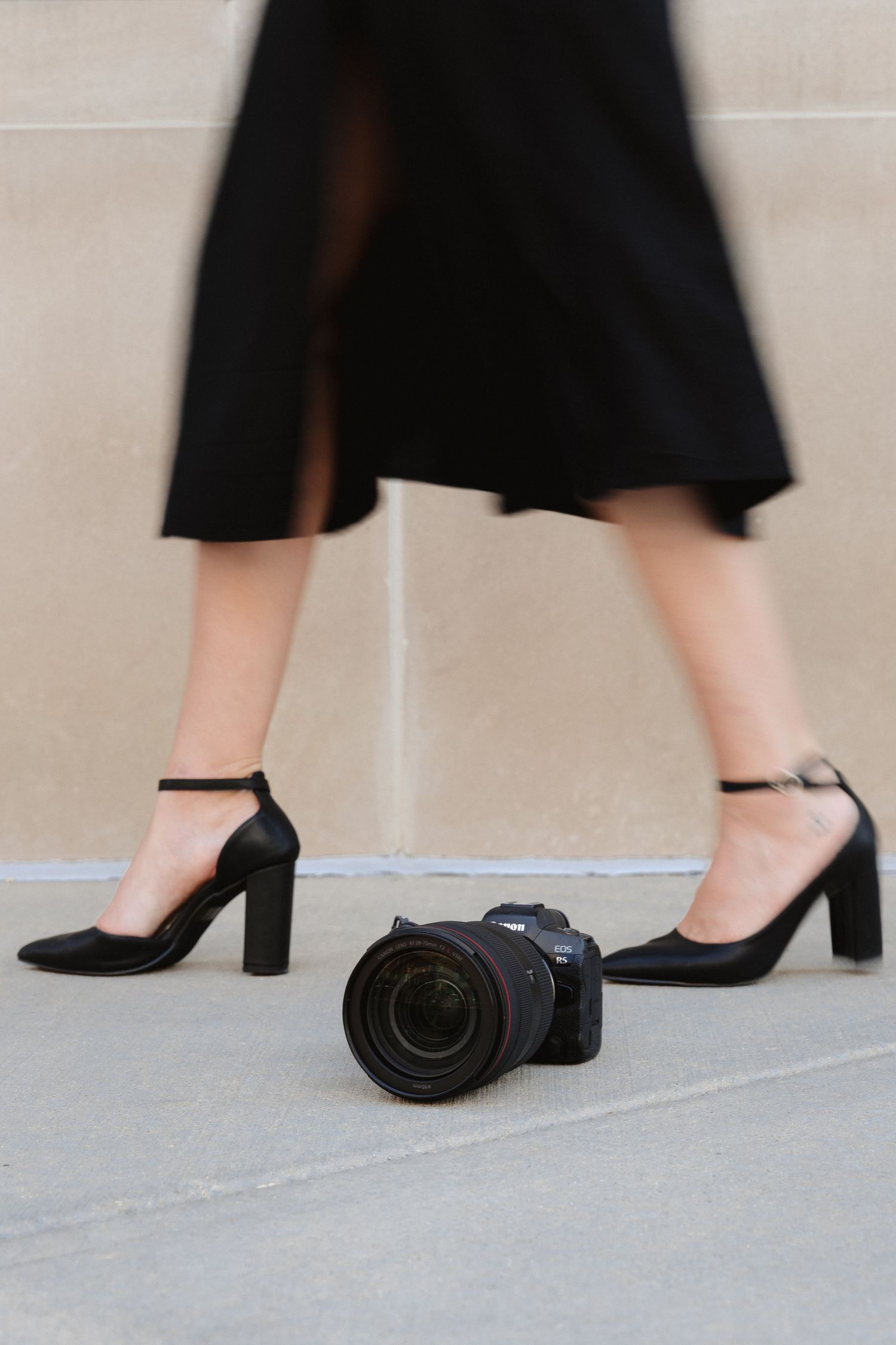 Woman in black heels and skirt standing beside a Canon DSLR camera placed on a sidewalk.