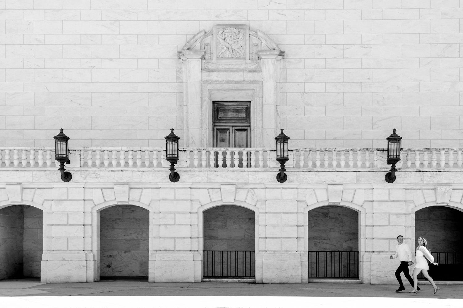 Two people walking past a grand neoclassical building facade with arched doorways and ornate lamp posts in black and white.