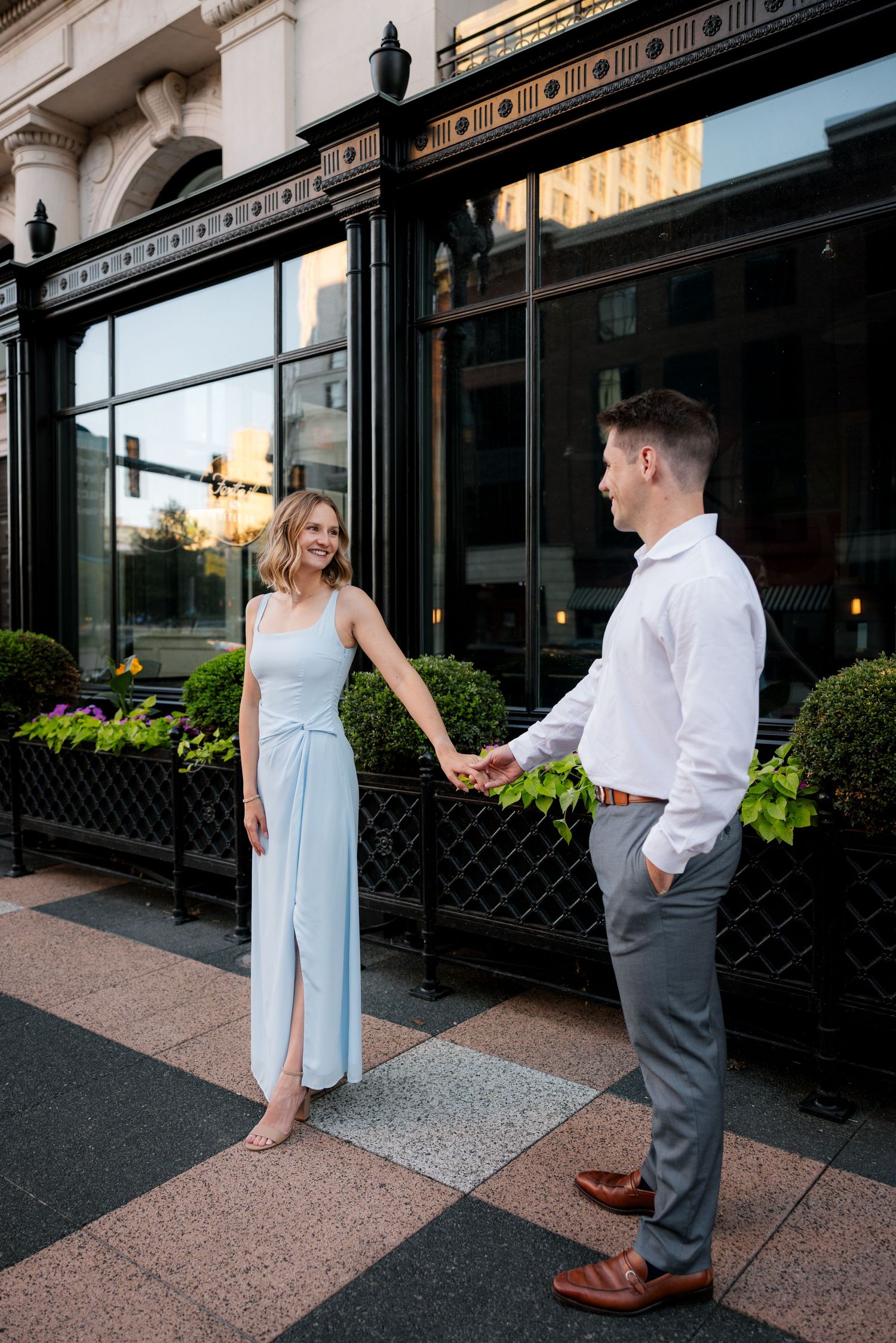 Couple holding hands and smiling outside a black-framed storefront with lush green planters.