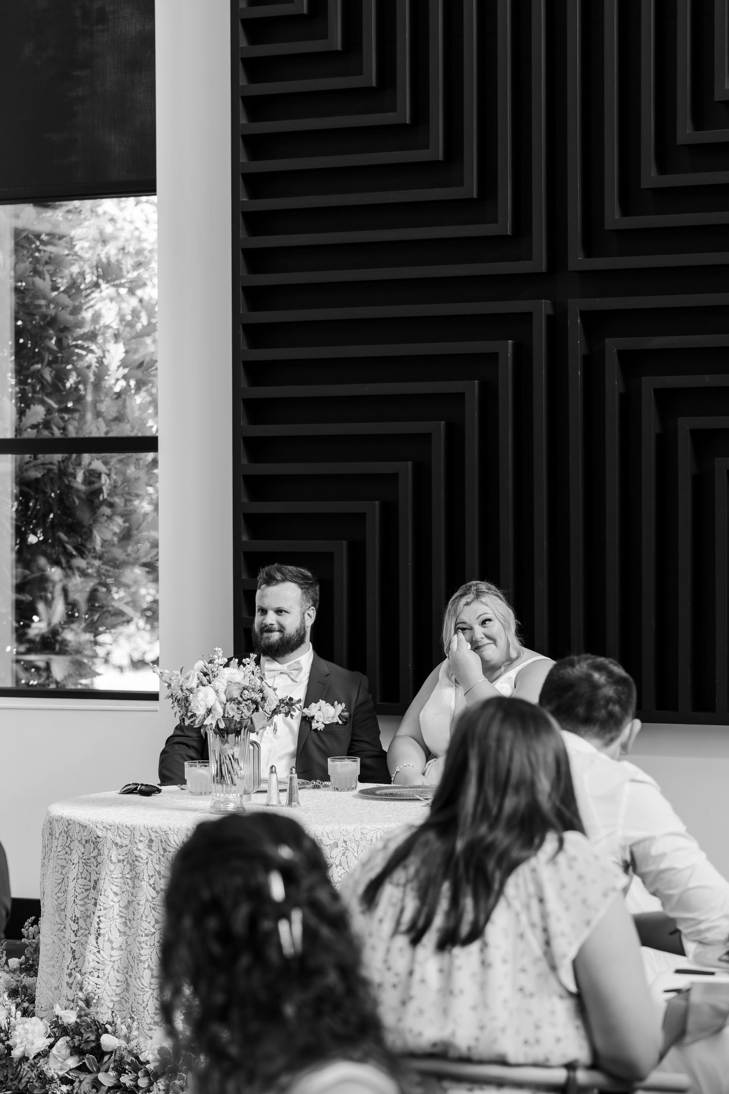 Black and white photo of a smiling bride and groom seated at a sweetheart table during their wedding reception.