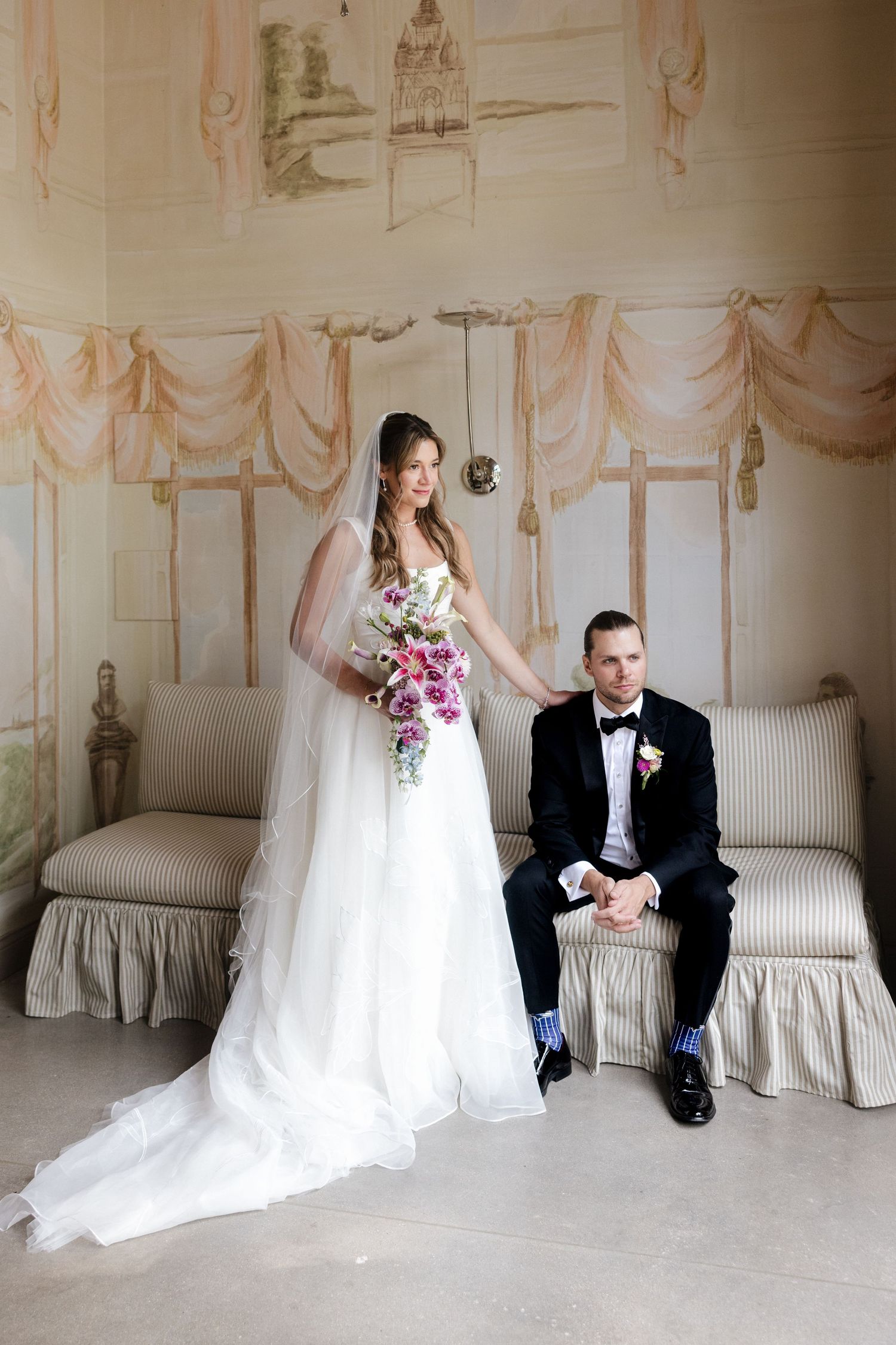 Bride in white gown holding floral bouquet stands beside seated groom in navy suit in elegant decorated room.