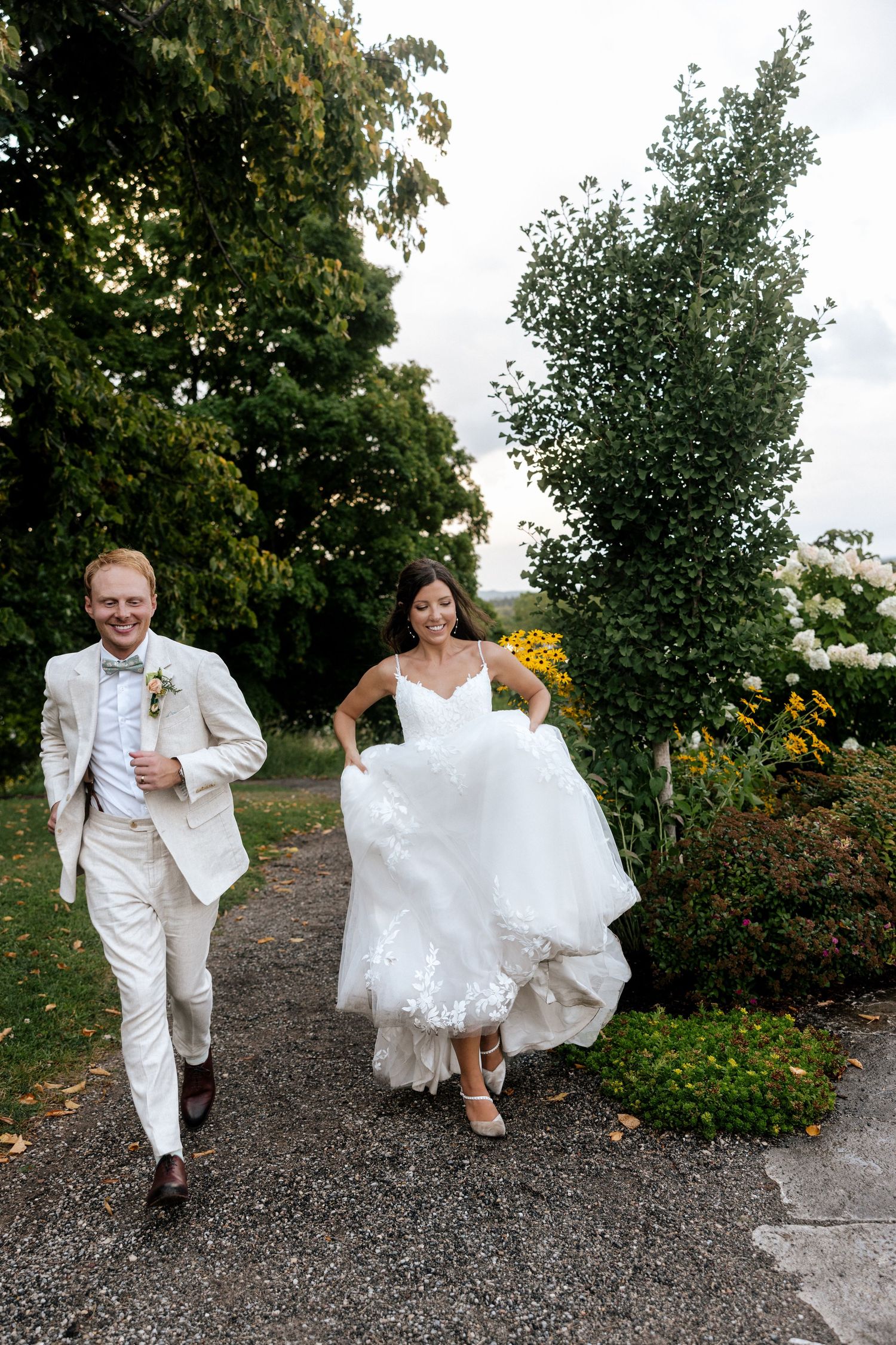 Smiling bride and groom walking through garden path, bride lifting lace wedding dress hem.