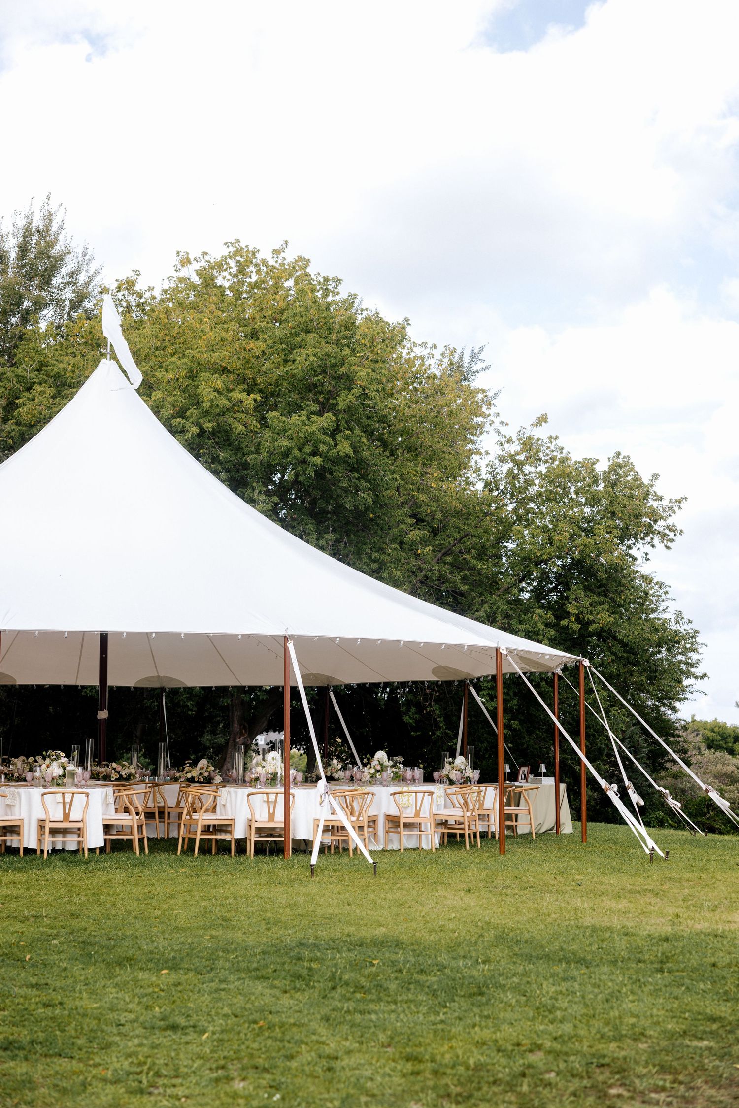 Elegant outdoor wedding reception under a large white sailcloth tent with floral centerpieces on round tables surrounded by greenery.