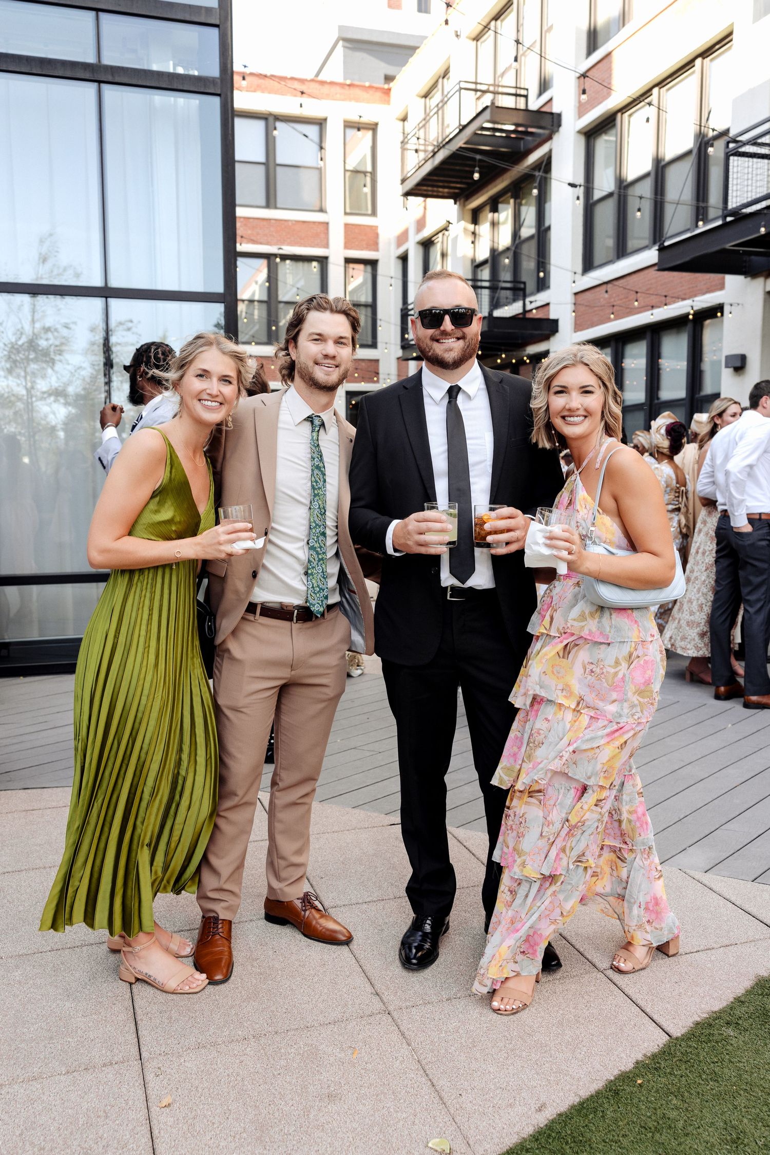 Four guests in elegant attire smile at an outdoor rooftop wedding reception, holding drinks on a sunny day.