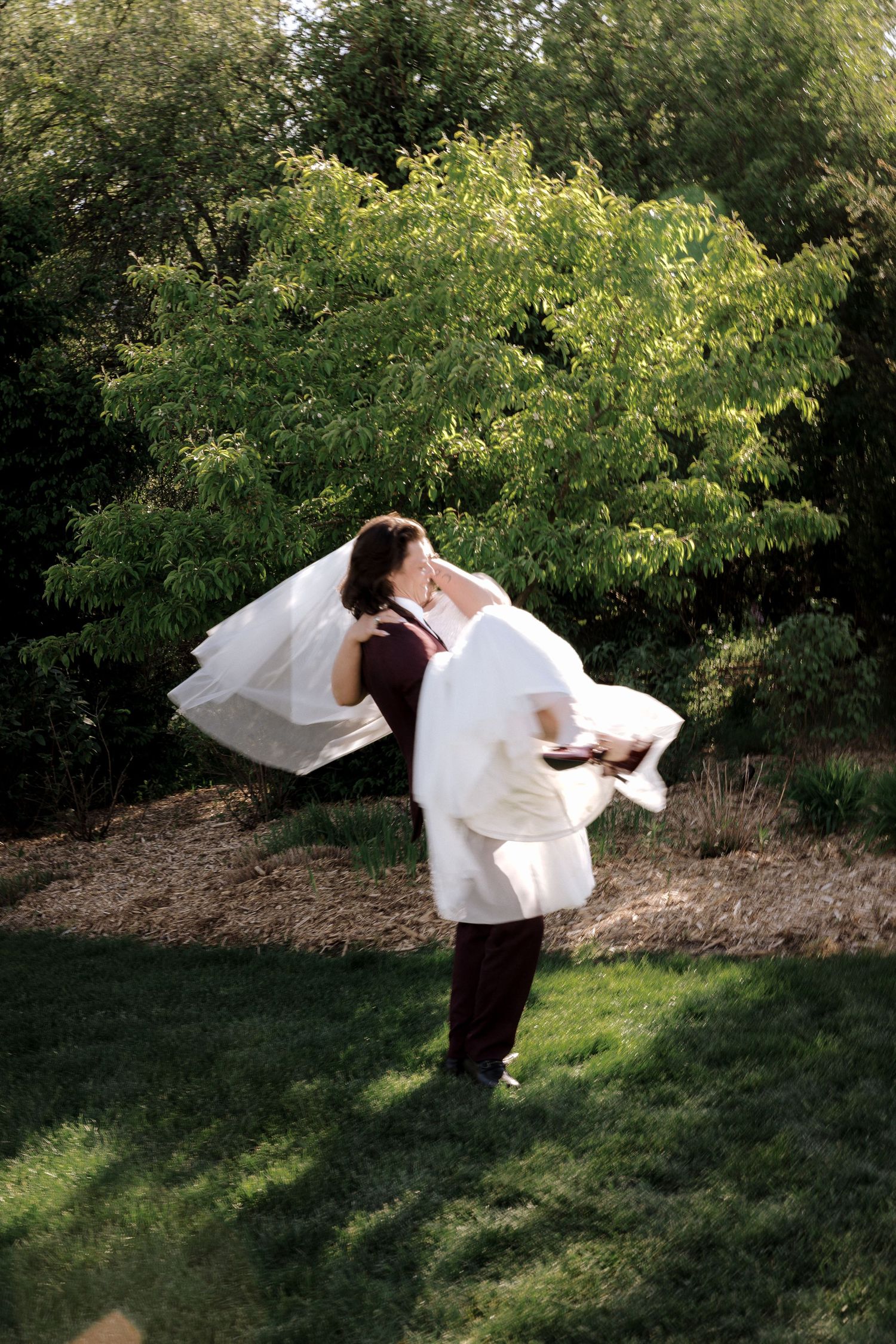 Groom in burgundy suit lifts bride in white gown and veil, spinning her outdoors amid lush green trees.