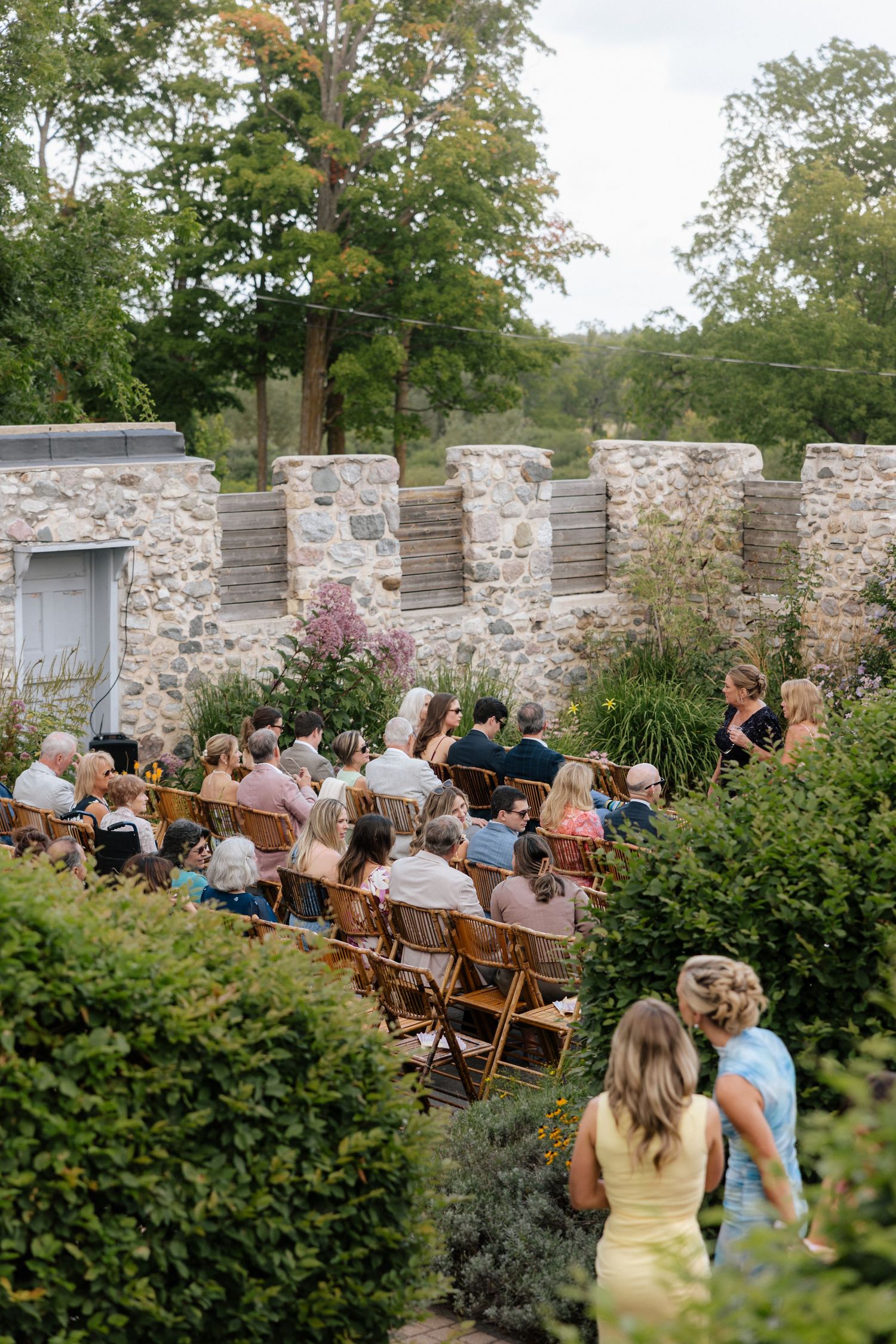 Outdoor wedding ceremony with guests seated on wooden chairs in a lush garden surrounded by stone walls and greenery.