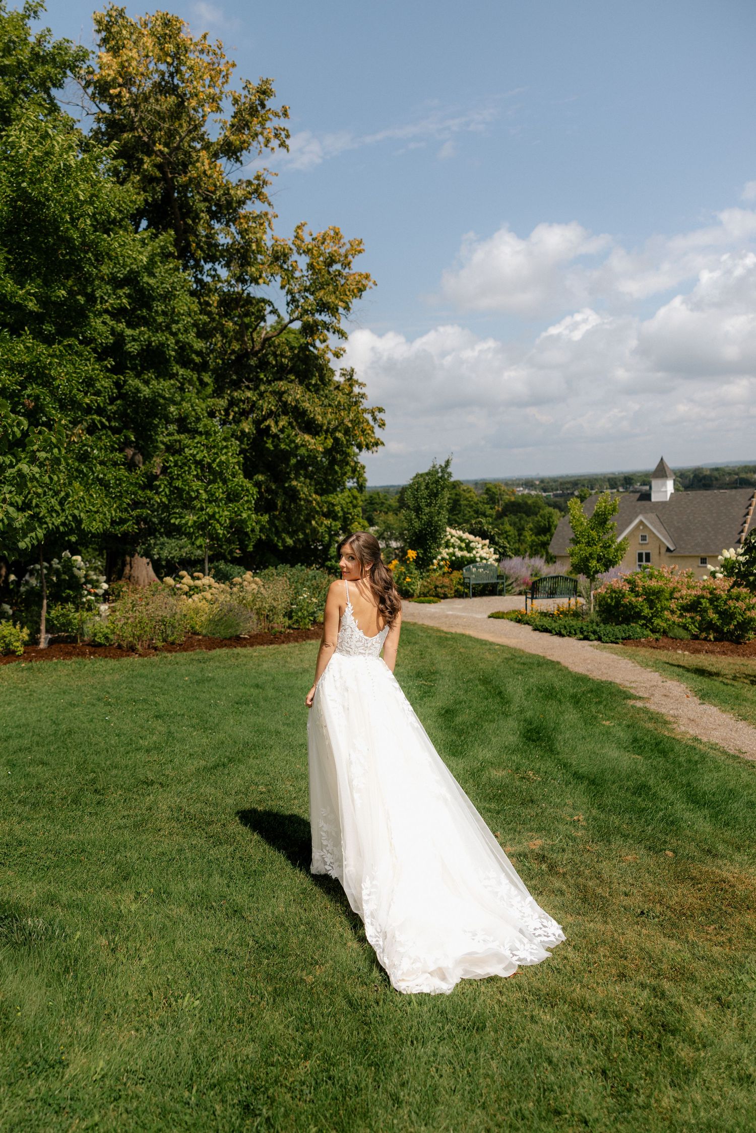 Bride in a backless lace wedding gown stands on lush green lawn surrounded by gardens and trees on a sunny day.