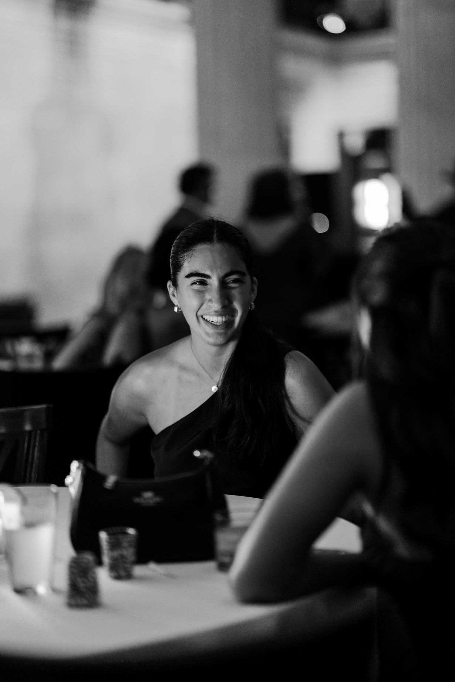 Black and white photo of a smiling woman in a one-shoulder dress at an elegant restaurant dinner event.