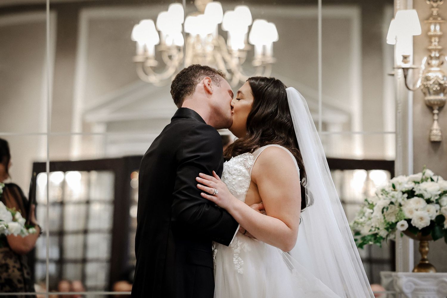 Bride and groom share romantic first kiss at altar with white floral arrangements in background.