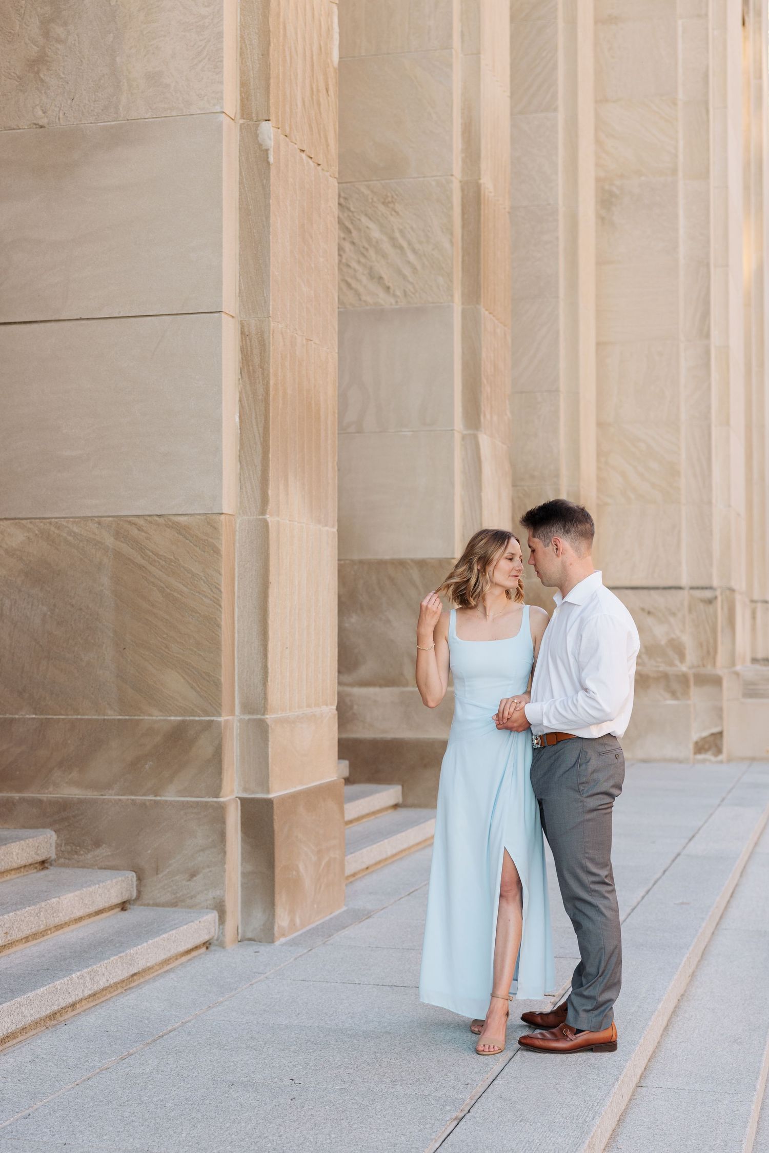 Couple embracing near stone columns, woman in light blue dress and man in white shirt and gray pants.