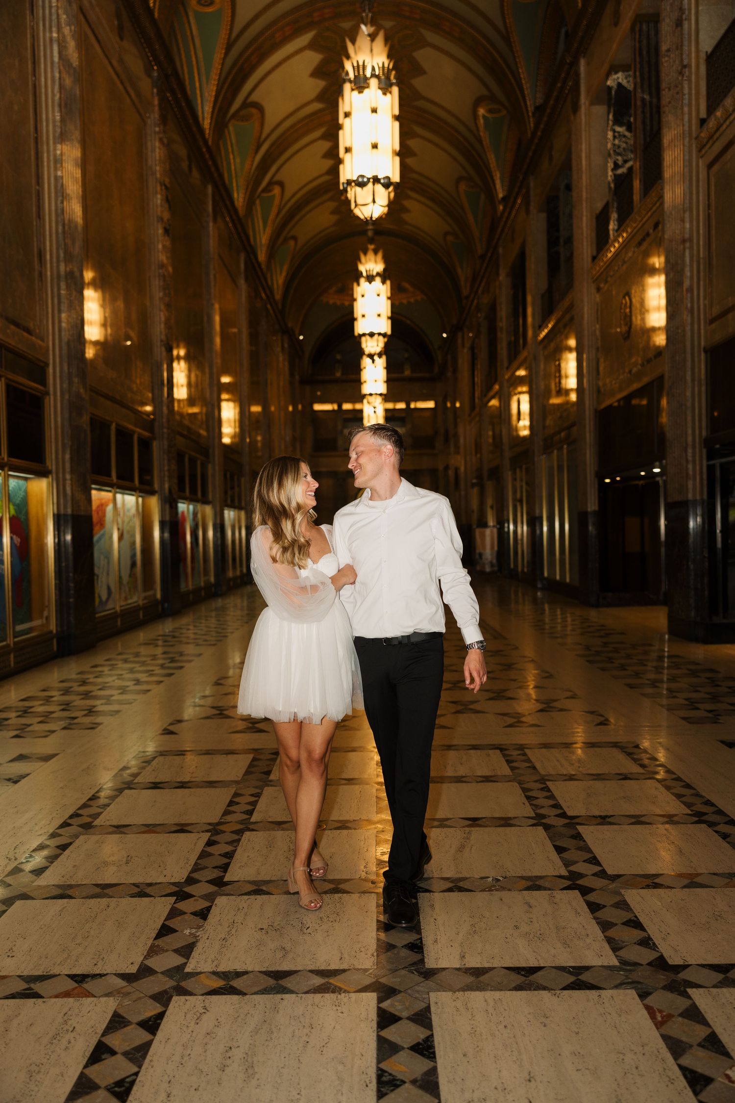 Couple walking hand-in-hand through an ornate golden hallway with arched ceilings and chandeliers.