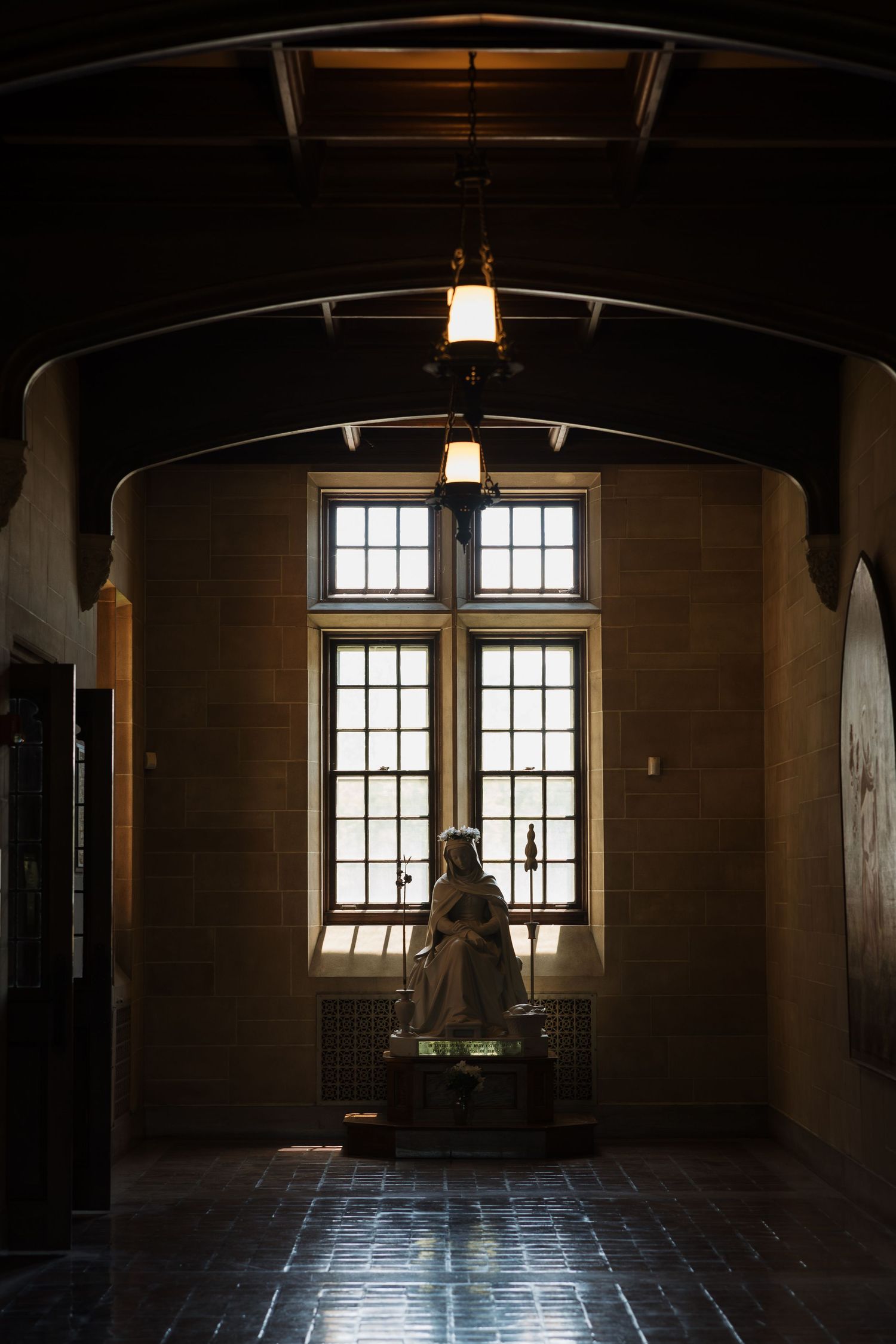 Dimly lit Gothic hall with arched ceiling, pendant lights, and a statue silhouetted against tall windows.