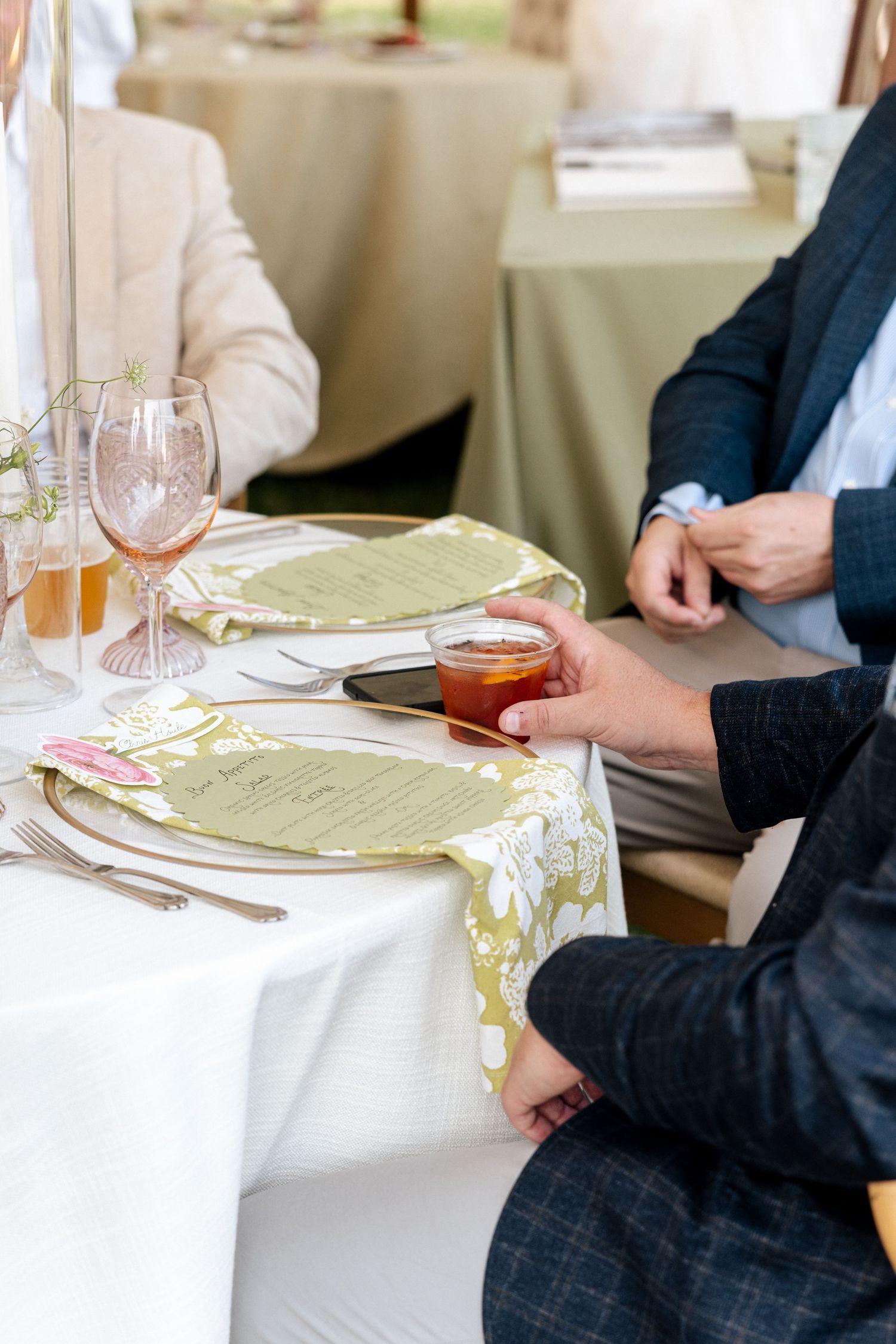 Elegant wedding reception table setting with green menus, floral napkins, pink glassware, and guests in formal attire.