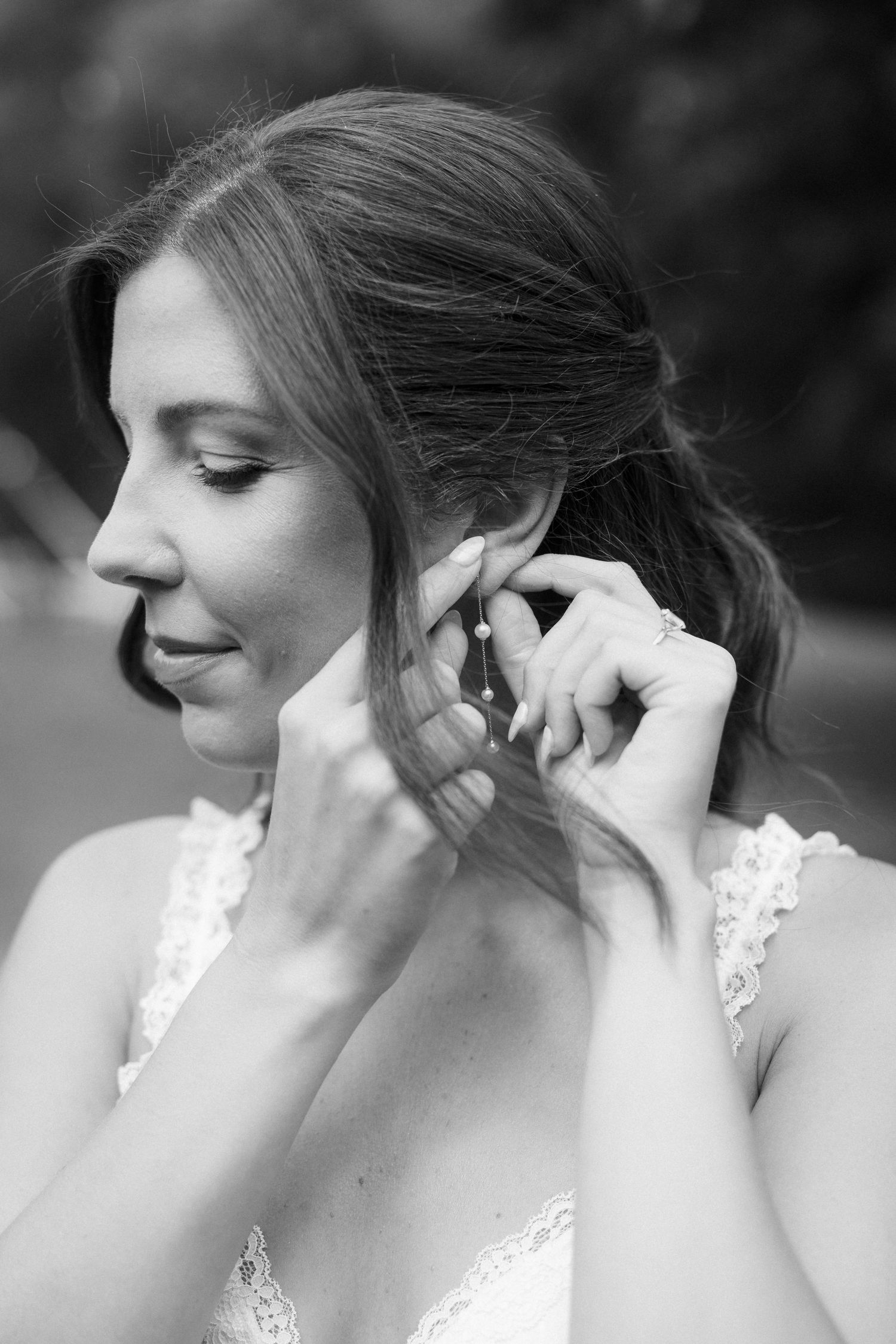 Black and white portrait of bride putting on earring in delicate lace wedding dress.