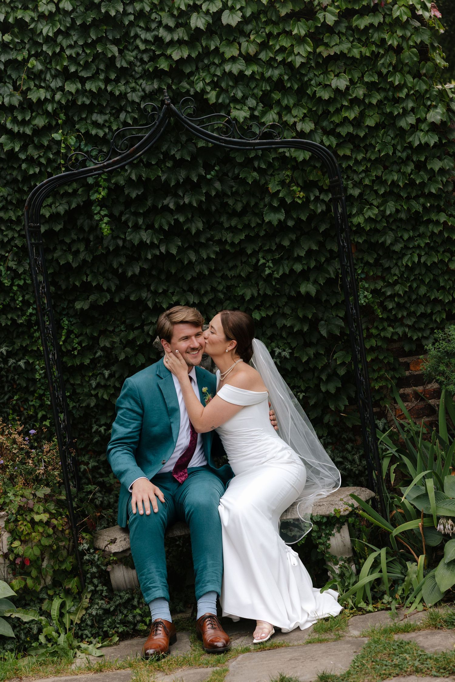 Bride in white gown kisses groom in teal suit on garden bench framed by lush ivy wall.