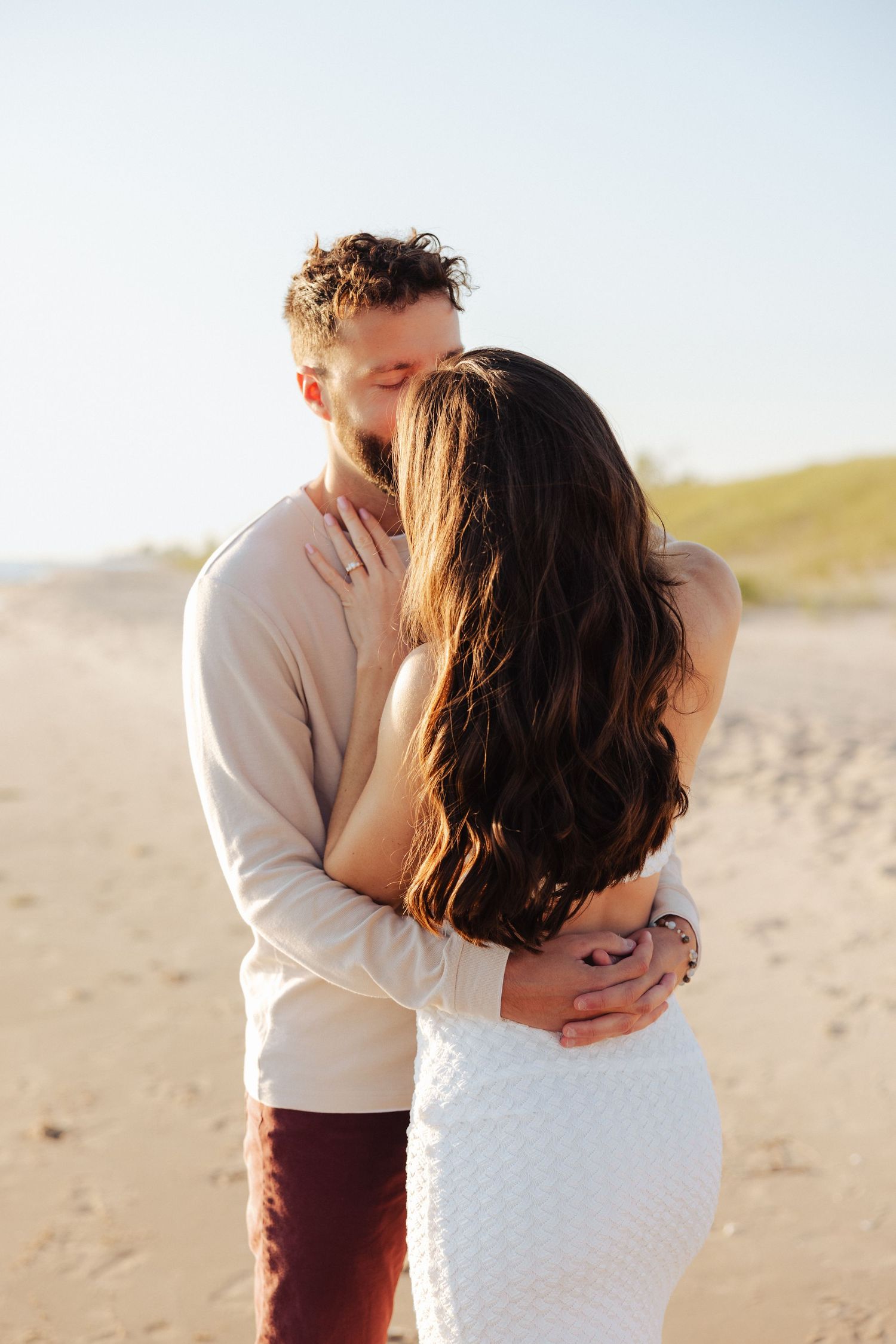 Couple embracing and kissing on a sandy beach at golden hour, woman in white dress with long dark hair.