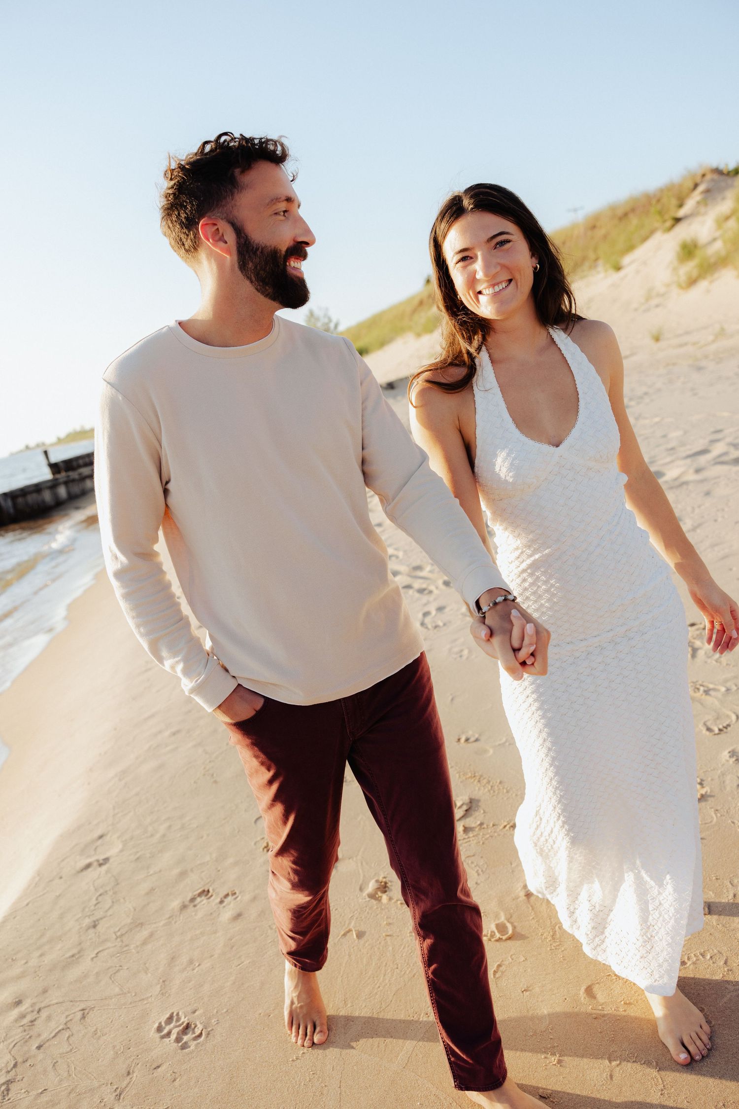 Happy couple walking barefoot on sandy beach, man in white shirt and burgundy pants, woman in white halter dress.