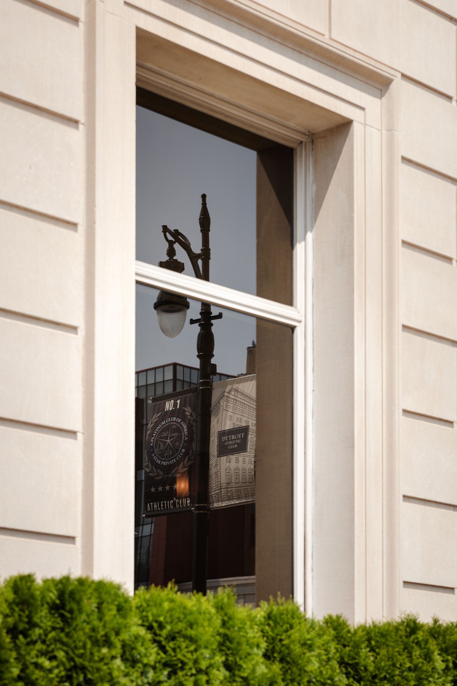 Neoclassical building window reflecting ornate street lamp and neighboring architecture with trimmed green hedges below.