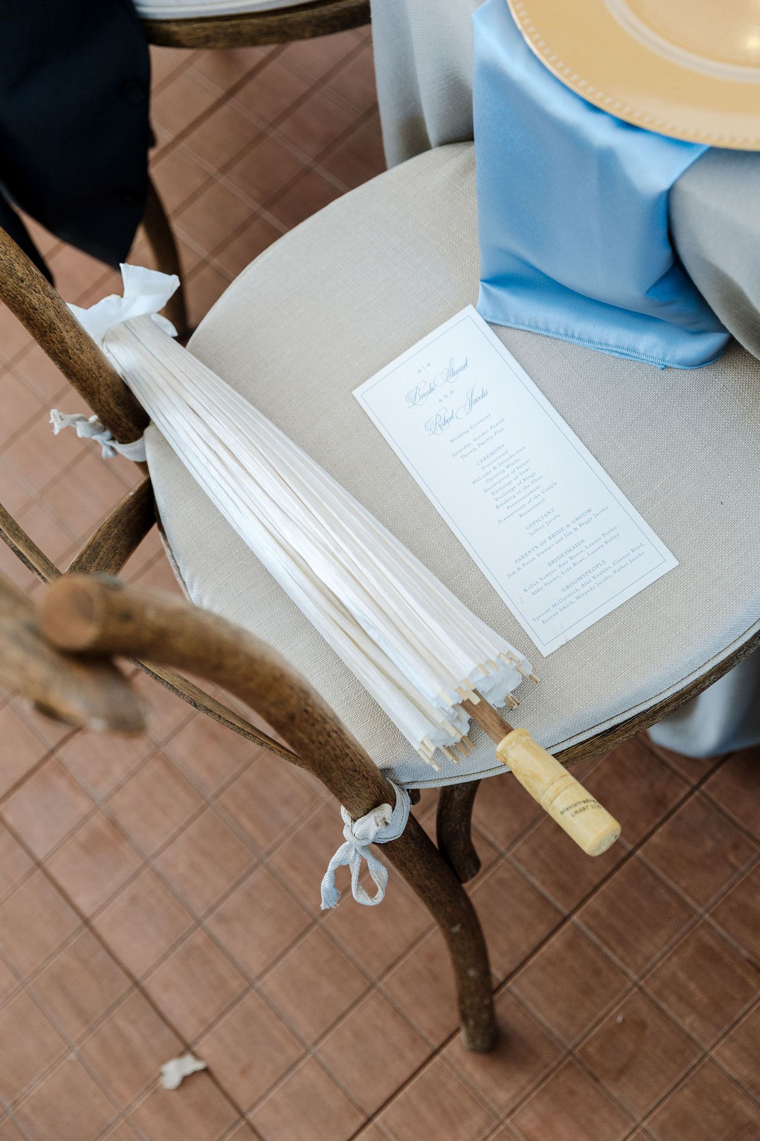 Wedding program and white parasol fan resting on a wooden chair at an elegant wedding reception venue.