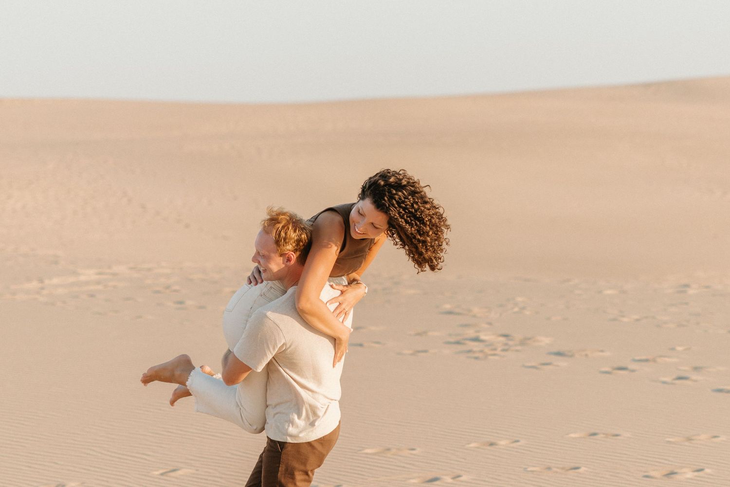 Man lifting woman in white dress on sandy desert dunes at sunset.