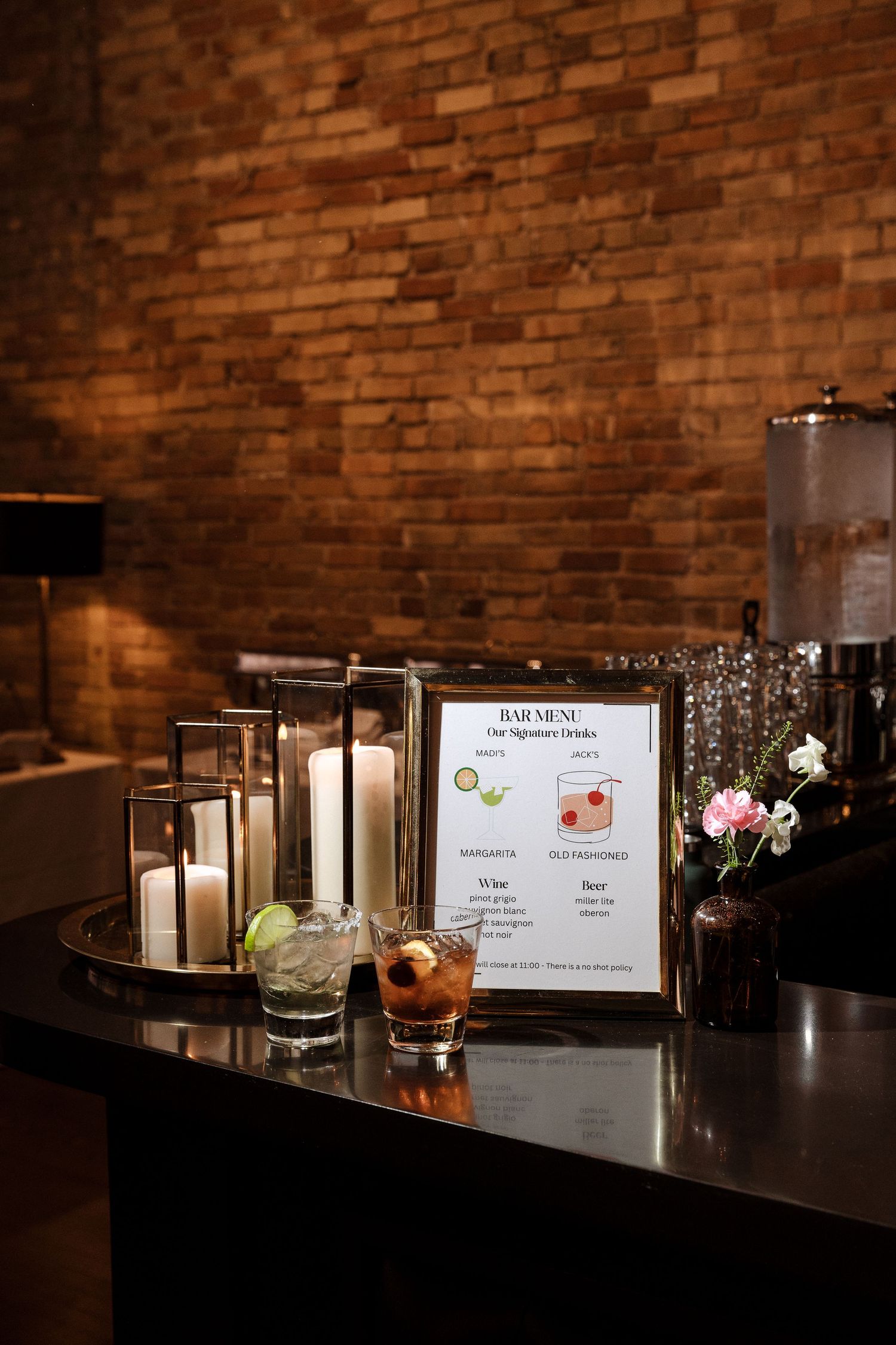 Elegant wedding bar setup with signature cocktail menu, candles, and fresh flowers on a dark counter against a brick wall.