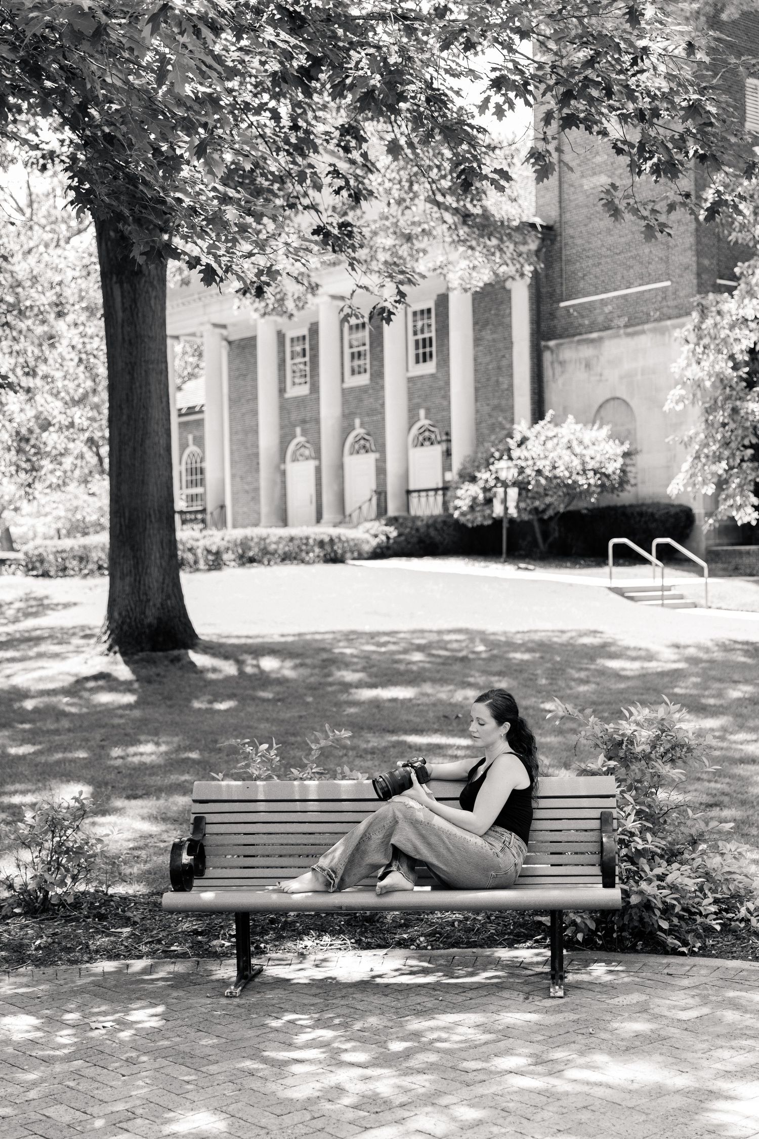 Young woman sitting on park bench under tree reading in black and white photo near brick building.