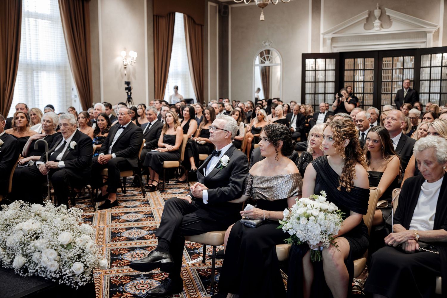Elegant wedding ceremony with guests seated in a grand ballroom adorned with white floral arrangements.
