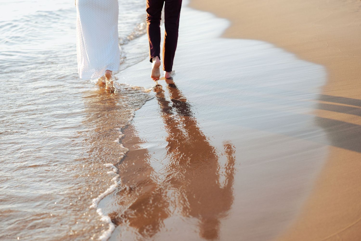 Barefoot couple in wedding attire walking along wet sandy beach shoreline with gentle waves.