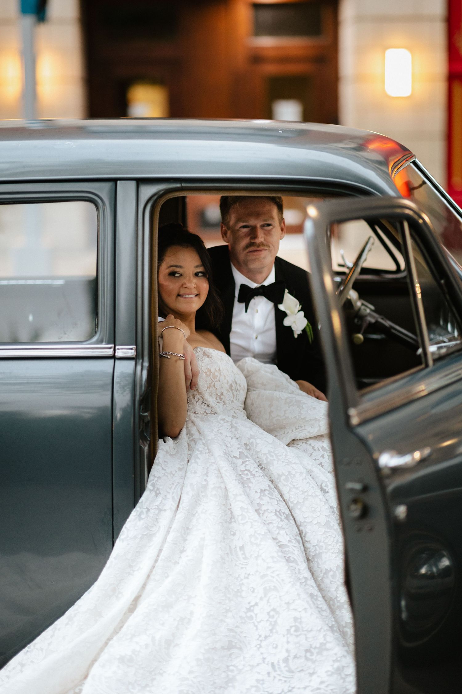Bride in lace gown and groom in tuxedo sitting together inside a vintage gray classic car on their wedding day.
