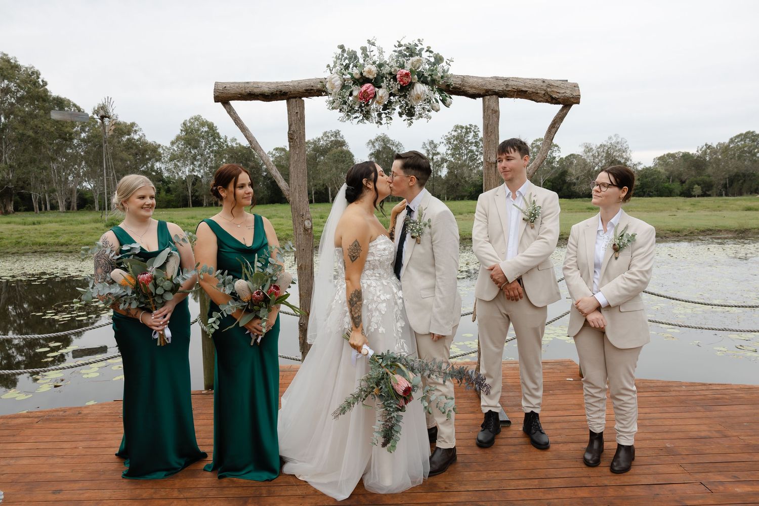 couple kissing in centre surrounded by bridal party. all standing on a jetty of a pond with green countryscape behind