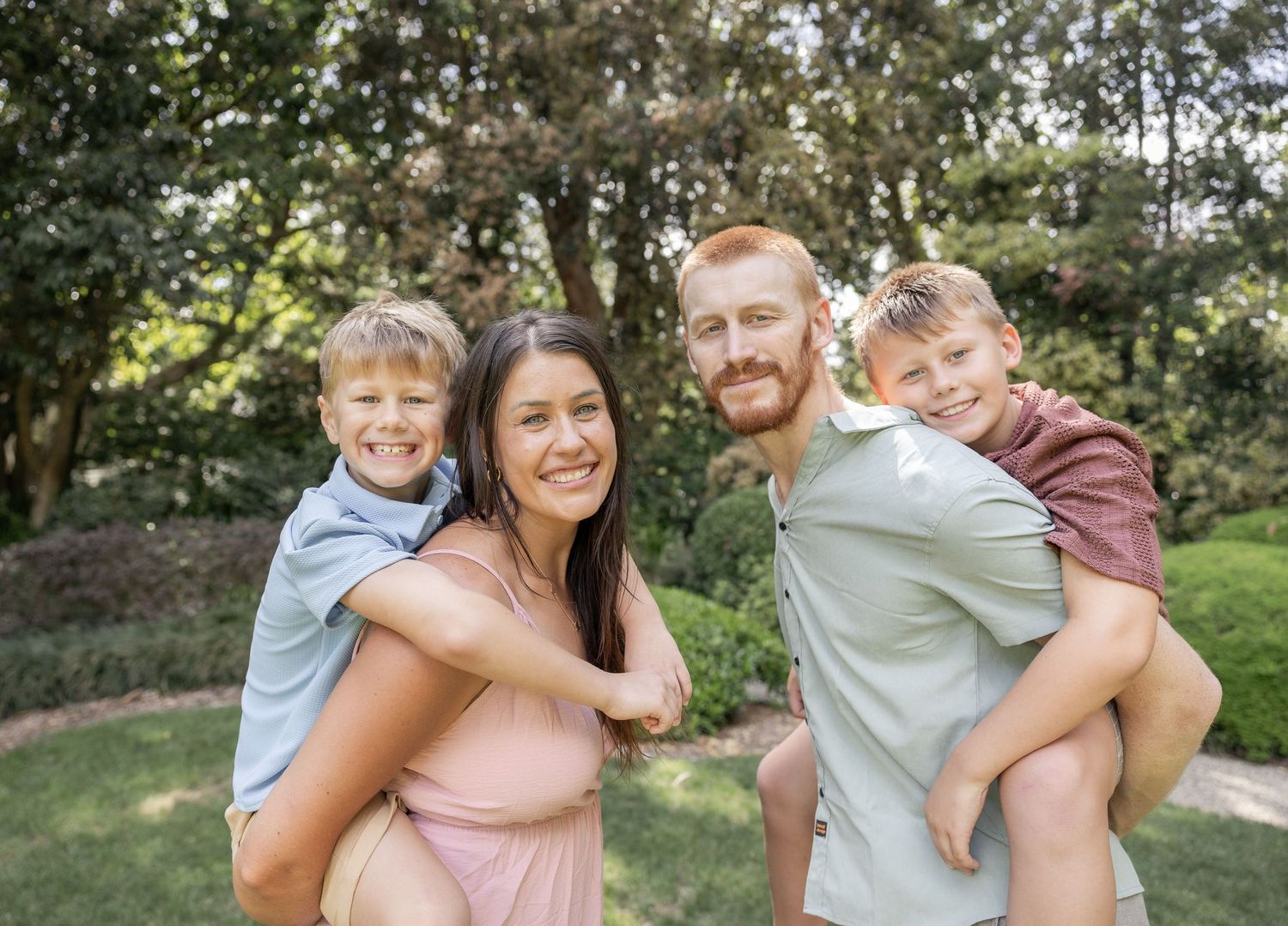 family of four, mum with boy on her back and dad with boy on his back all smiling at camera. natural greenscape background