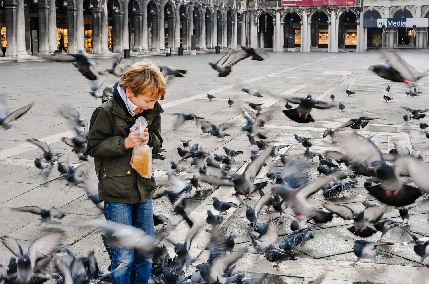 Young child surrounded by a flock of pigeons taking flight in a historic European plaza while holding a bag.