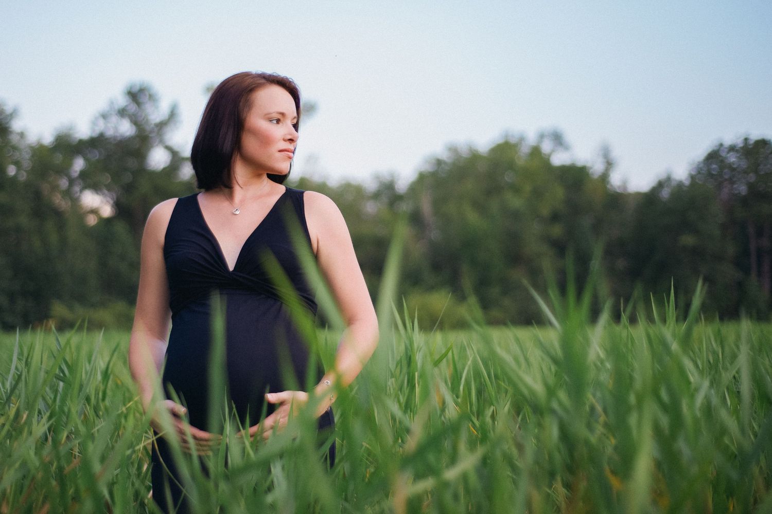 Woman in black dress standing in tall green grass field with trees in background.