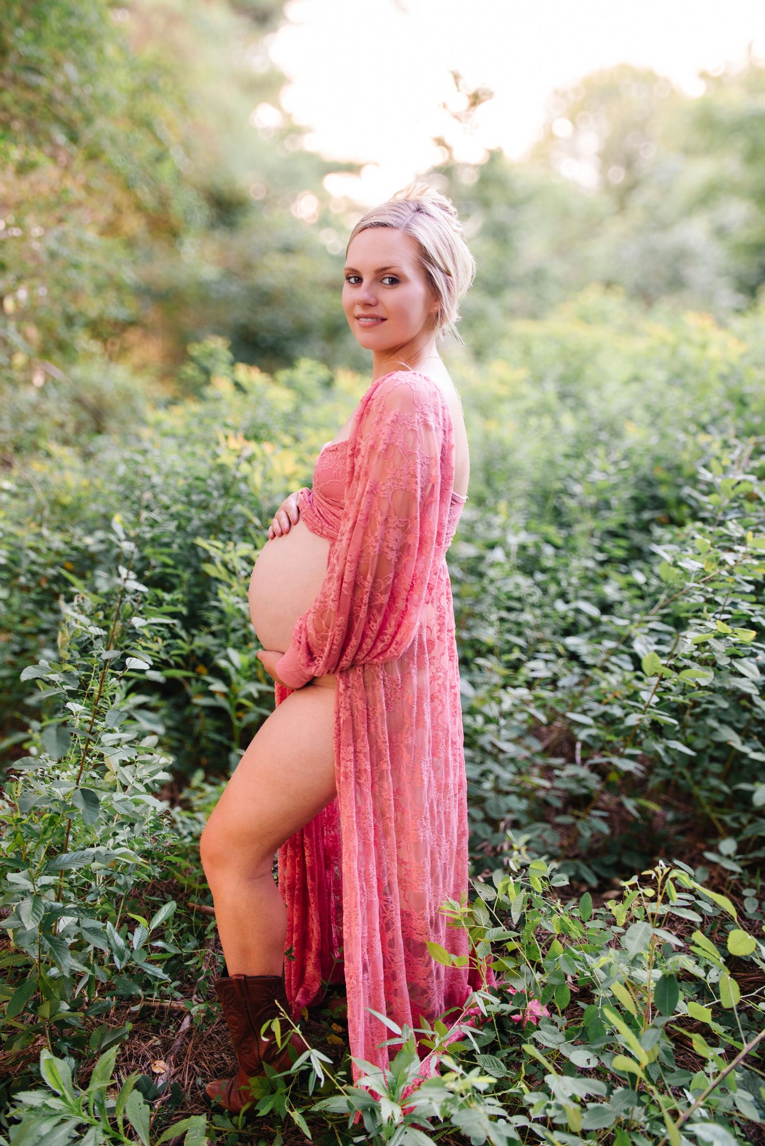 Pregnant woman in pink floral sheer robe cradling baby bump outdoors in lush green garden during golden hour.