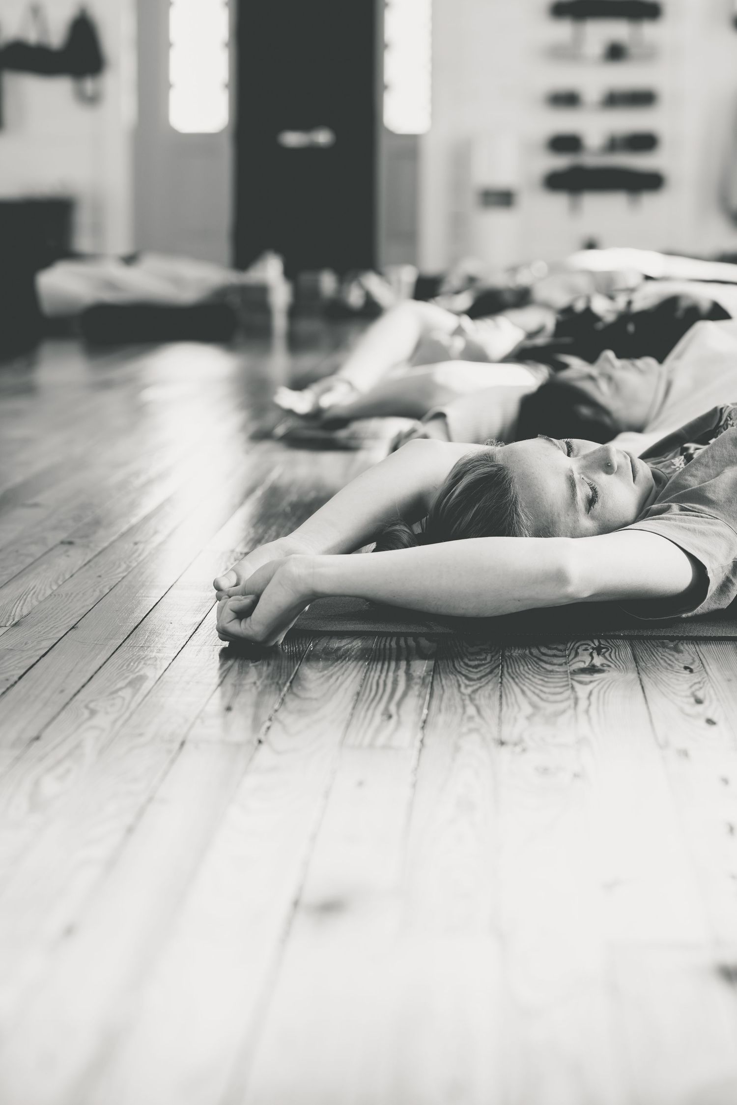 Black and white photo of yoga students lying in savasana pose on wooden studio floor.