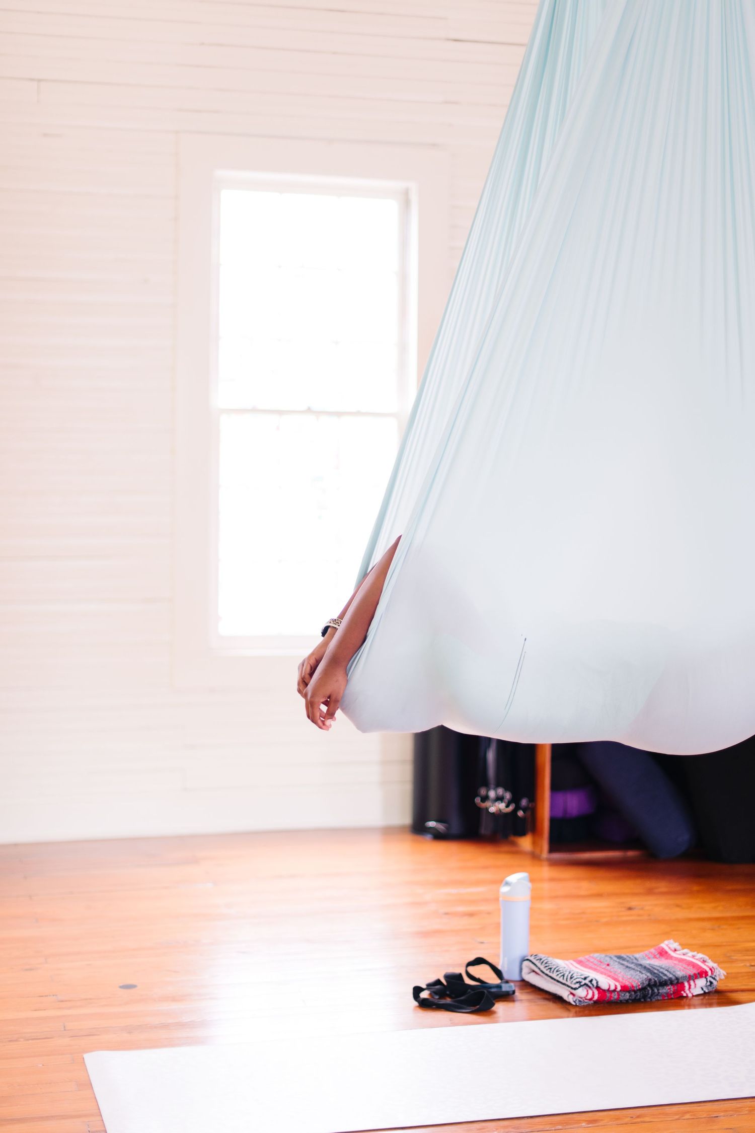 Person wrapped in a light blue aerial yoga silk hammock beside a bright window in a white studio.