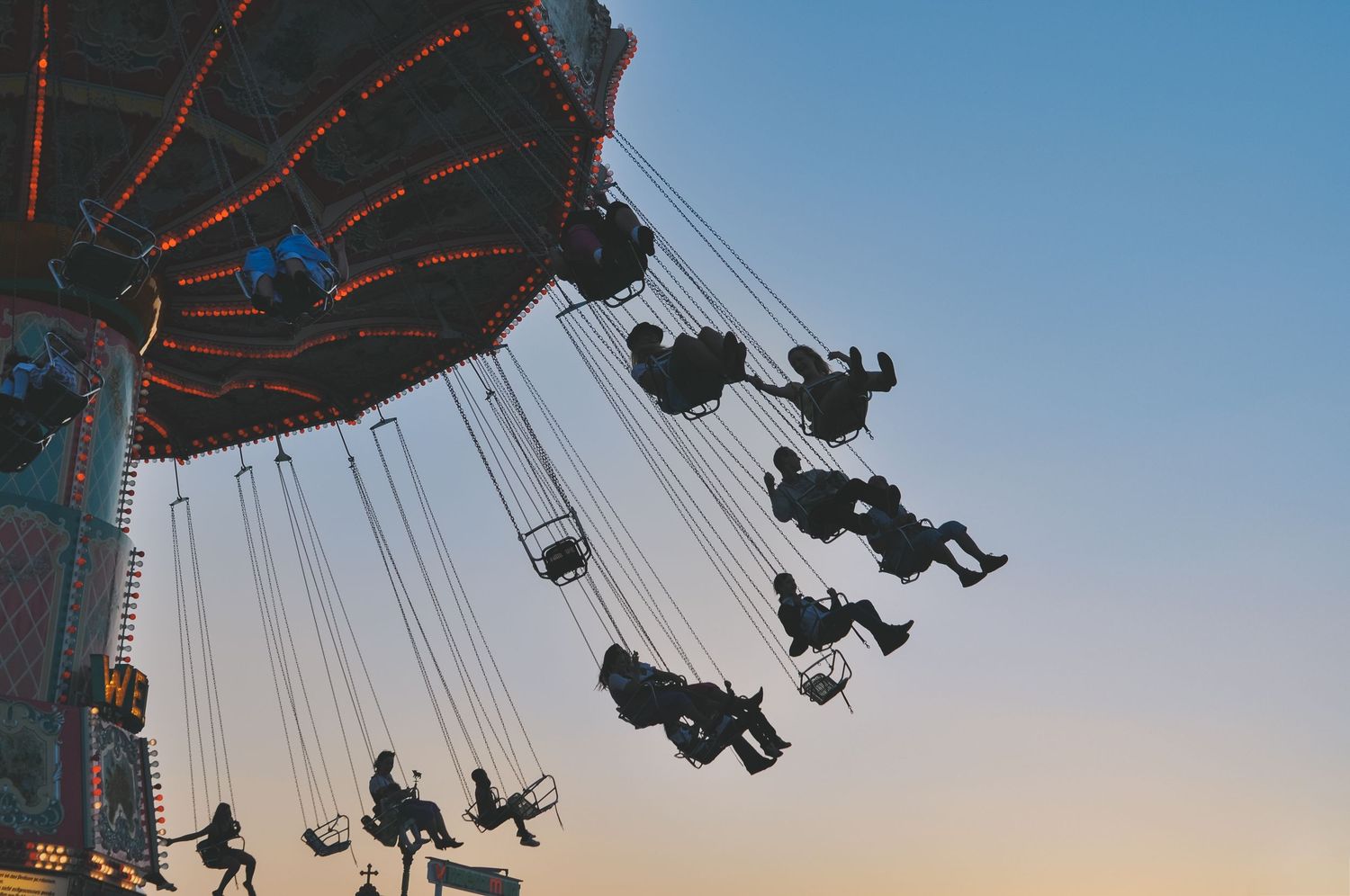 Silhouettes of people riding a chain swing carousel at dusk against a colorful sunset sky at an amusement park.