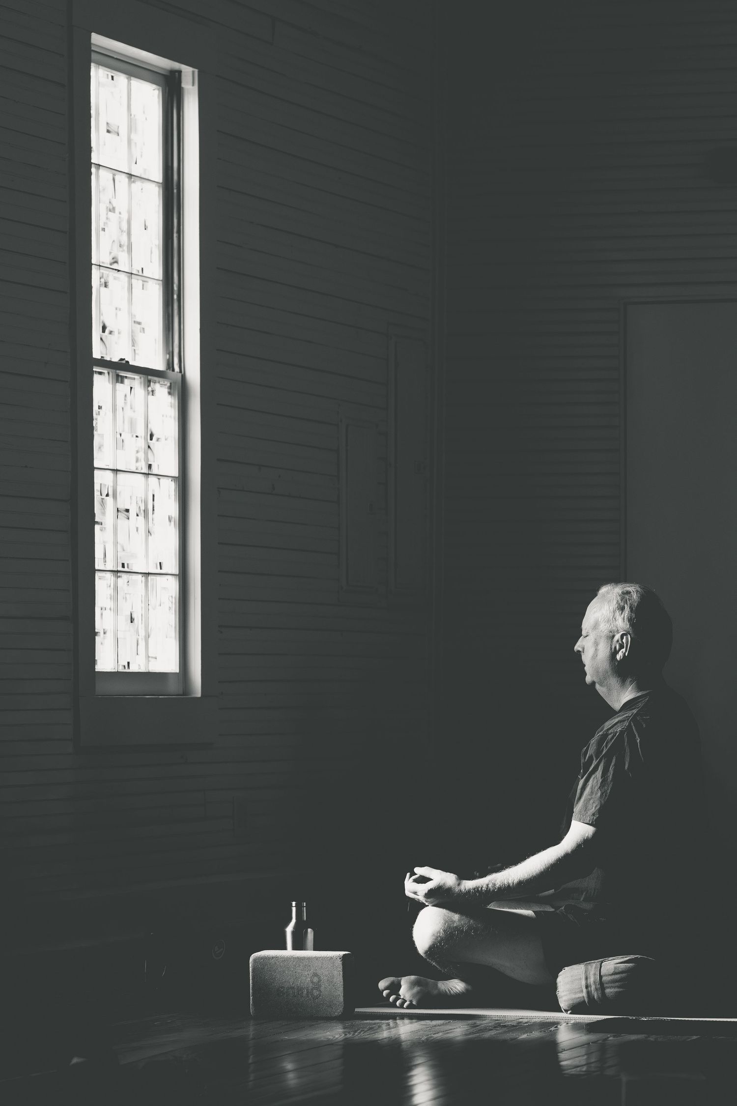 Black and white photo of man meditating in lotus pose by window in dark room.