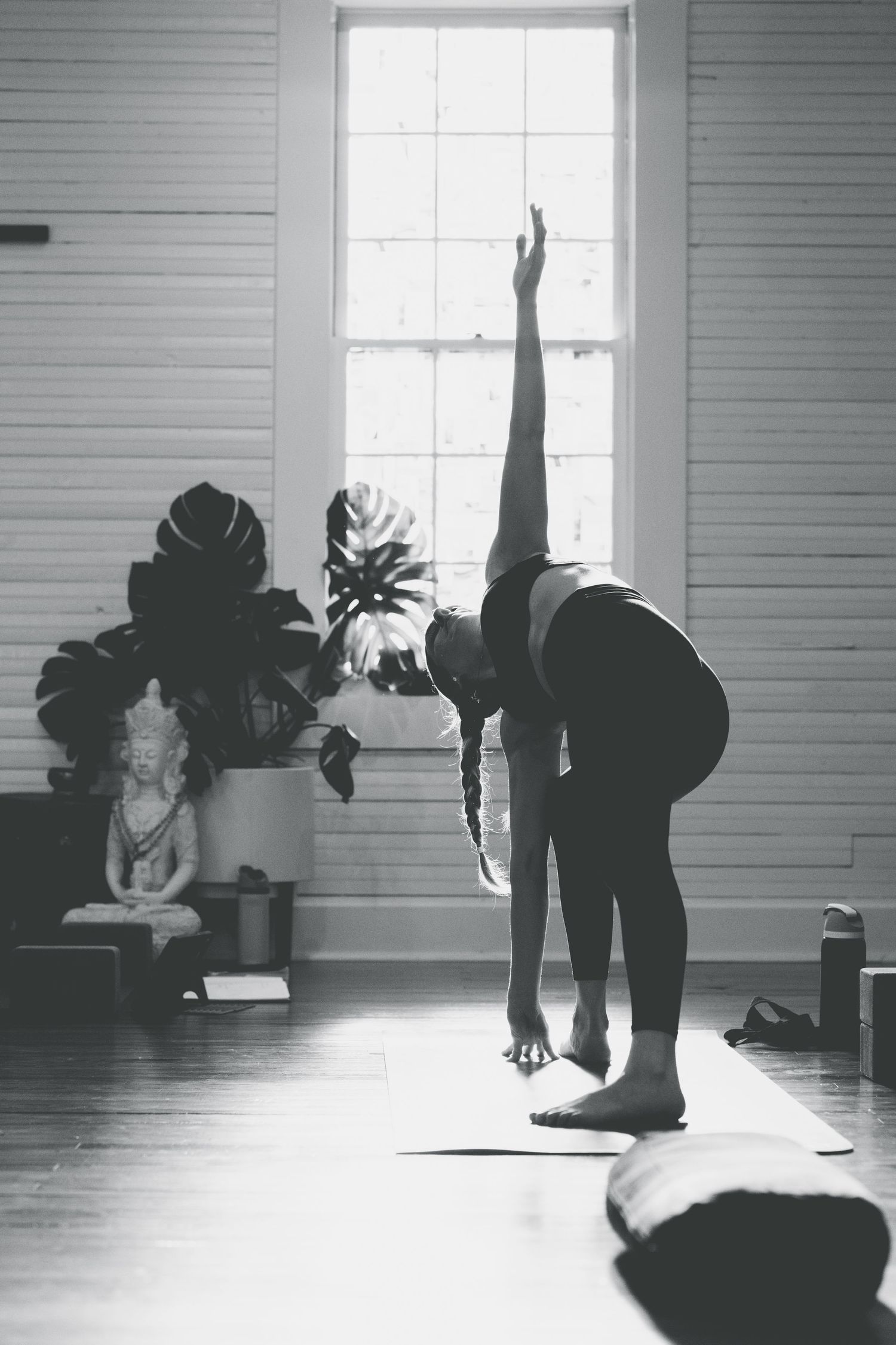 Woman practicing yoga in a sunlit studio, performing a standing forward bend with one arm raised toward the ceiling.