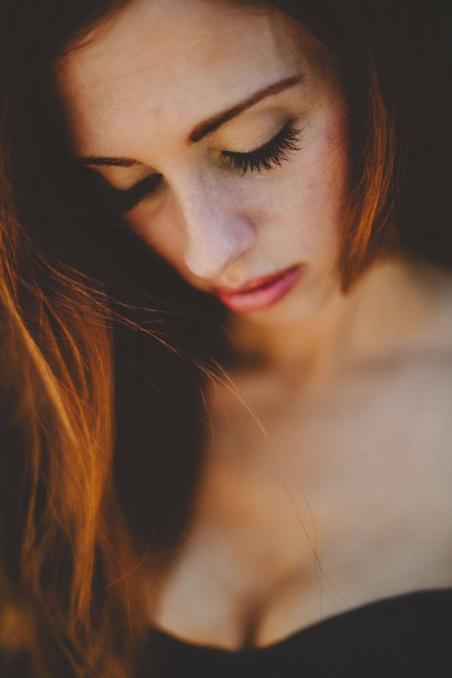Close-up portrait of a freckled redhead woman with long lashes looking down in warm moody lighting.