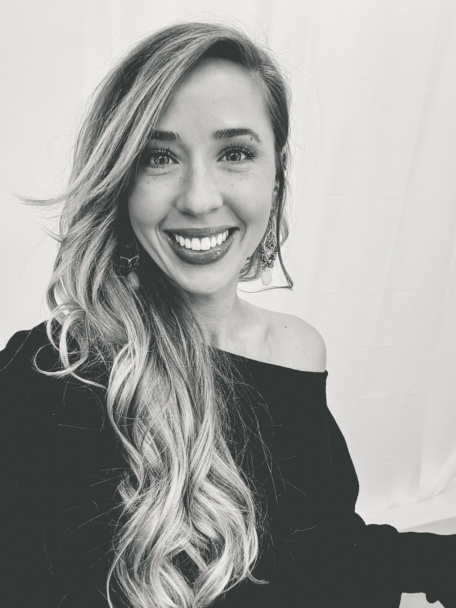 Smiling young woman with long wavy blonde hair wearing black off-shoulder top and statement earrings in black and white photo.