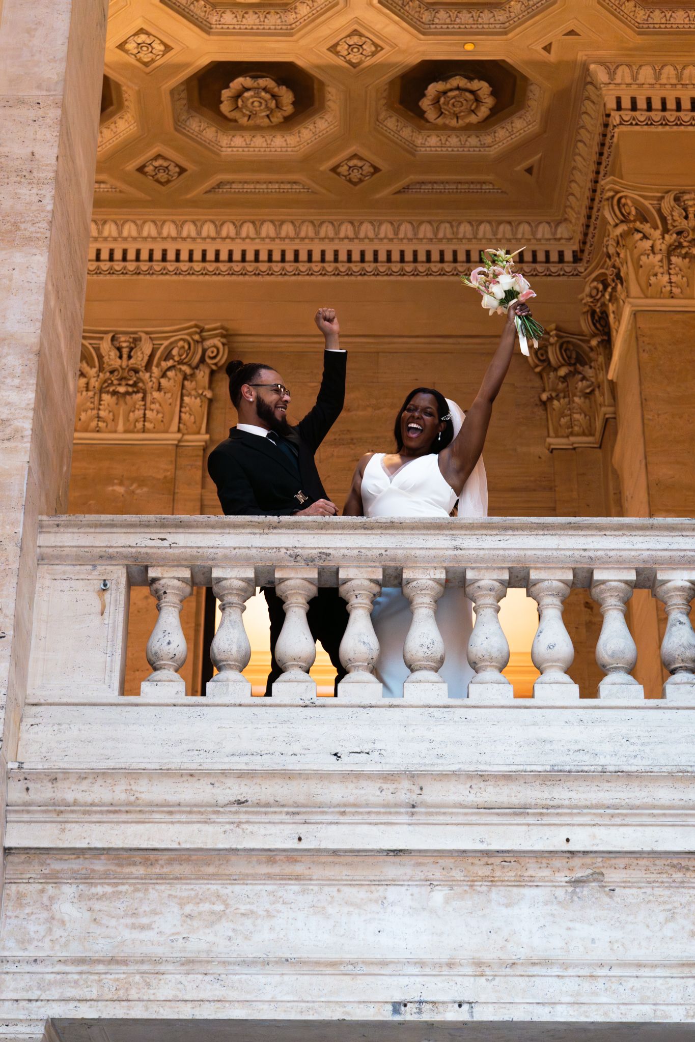 Joyful newlywed couple celebrating on ornate marble balcony with arms raised in elegant wedding venue.