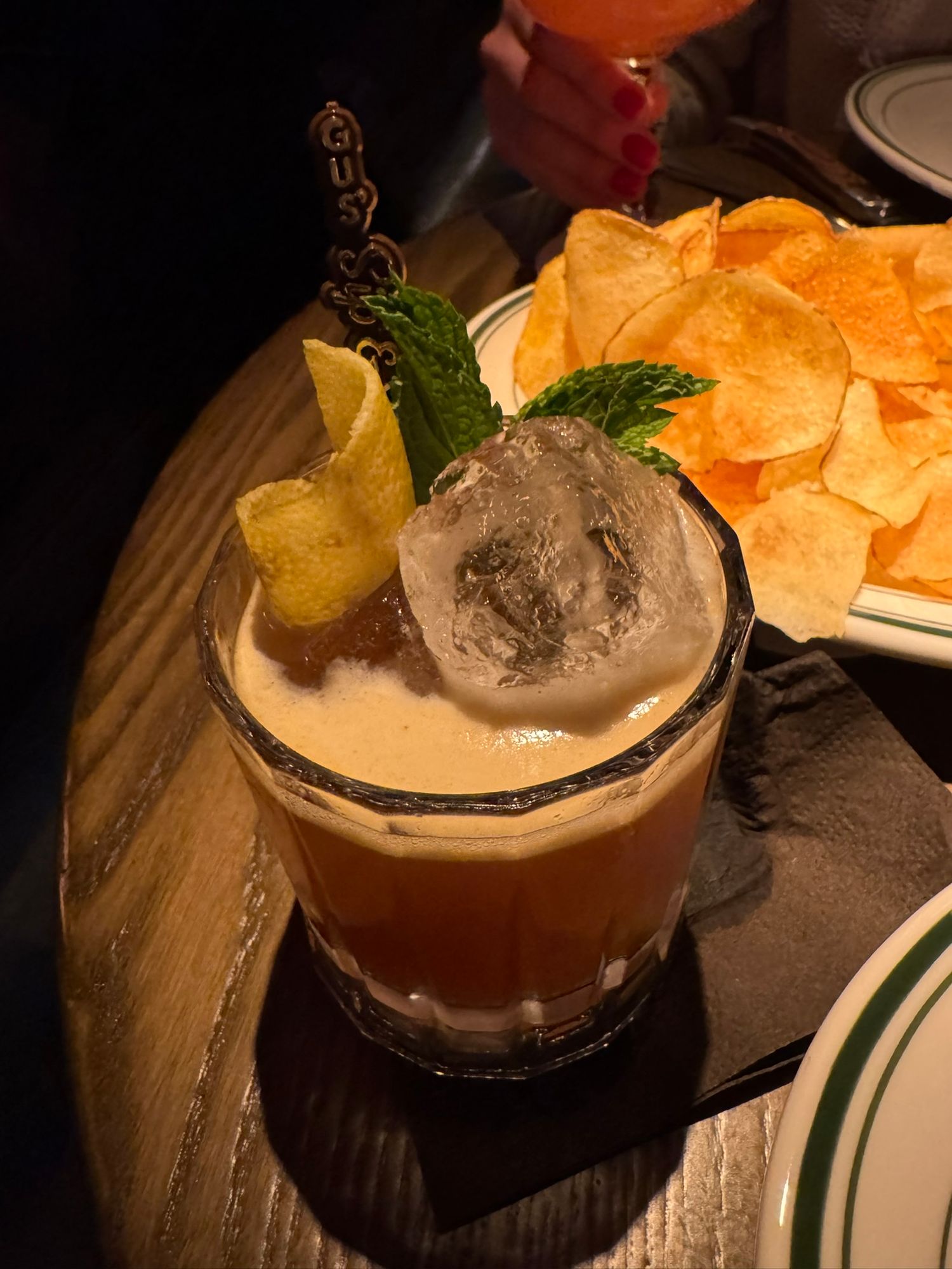 Craft cocktail with large ice sphere, mint garnish, and pineapple slice served alongside crispy chips on a wooden table | Sydney Allen Photo