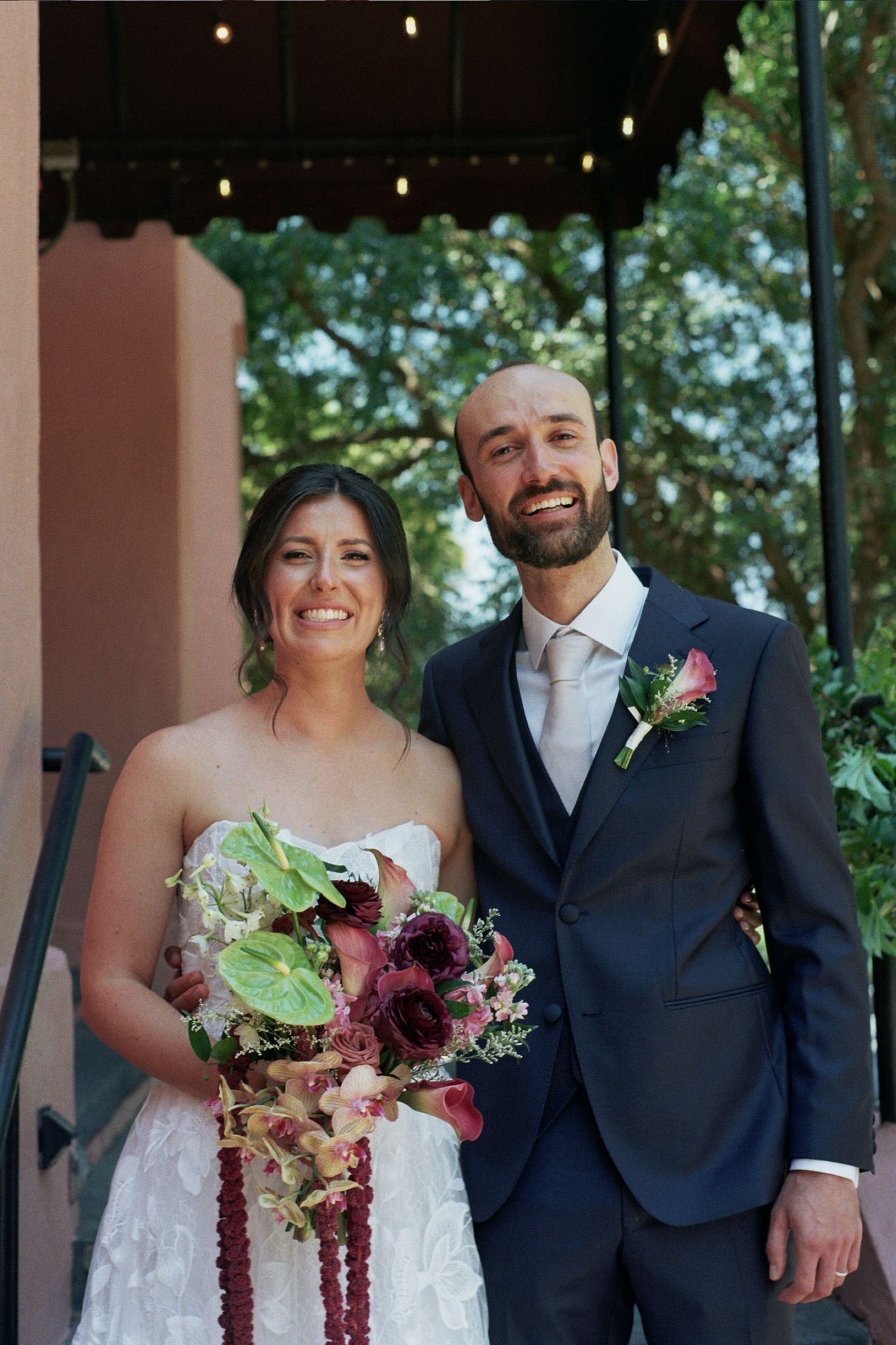 Smiling bride and groom pose together outdoors, she holds a burgundy bouquet, he wears a navy suit with floral boutonniere.