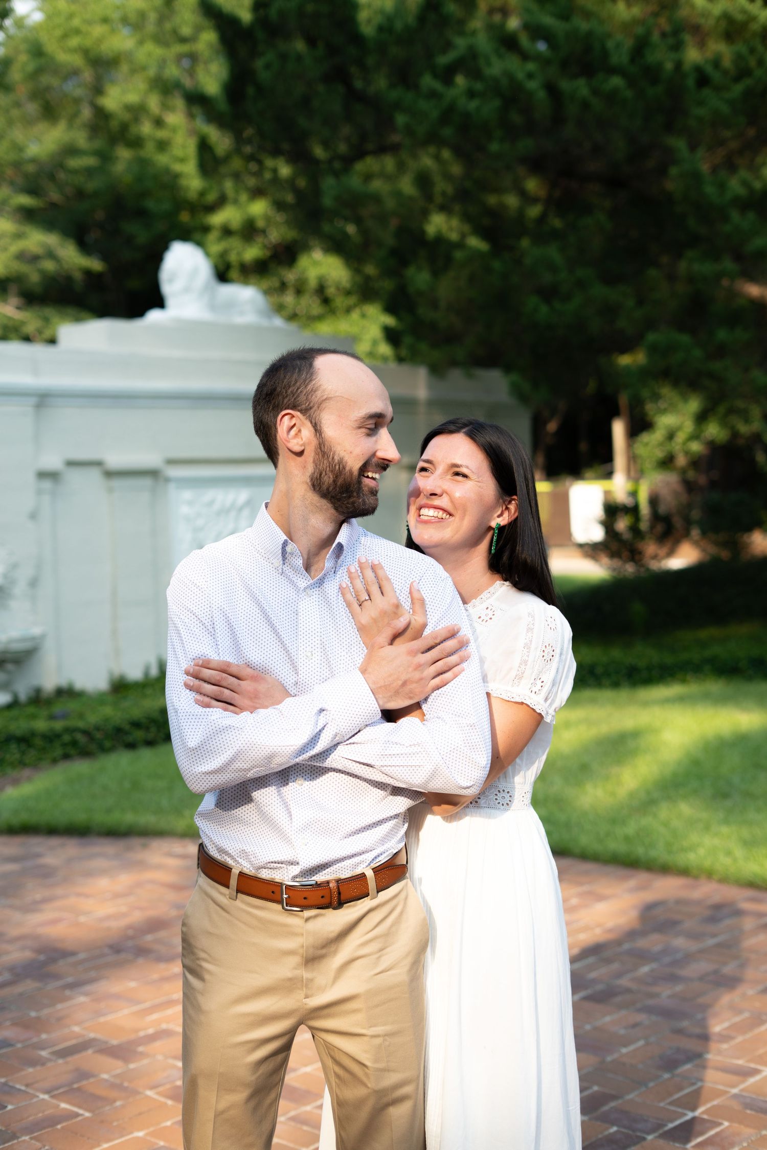 A smiling couple embraces outdoors, woman in white dress showing engagement ring, lush green garden background.