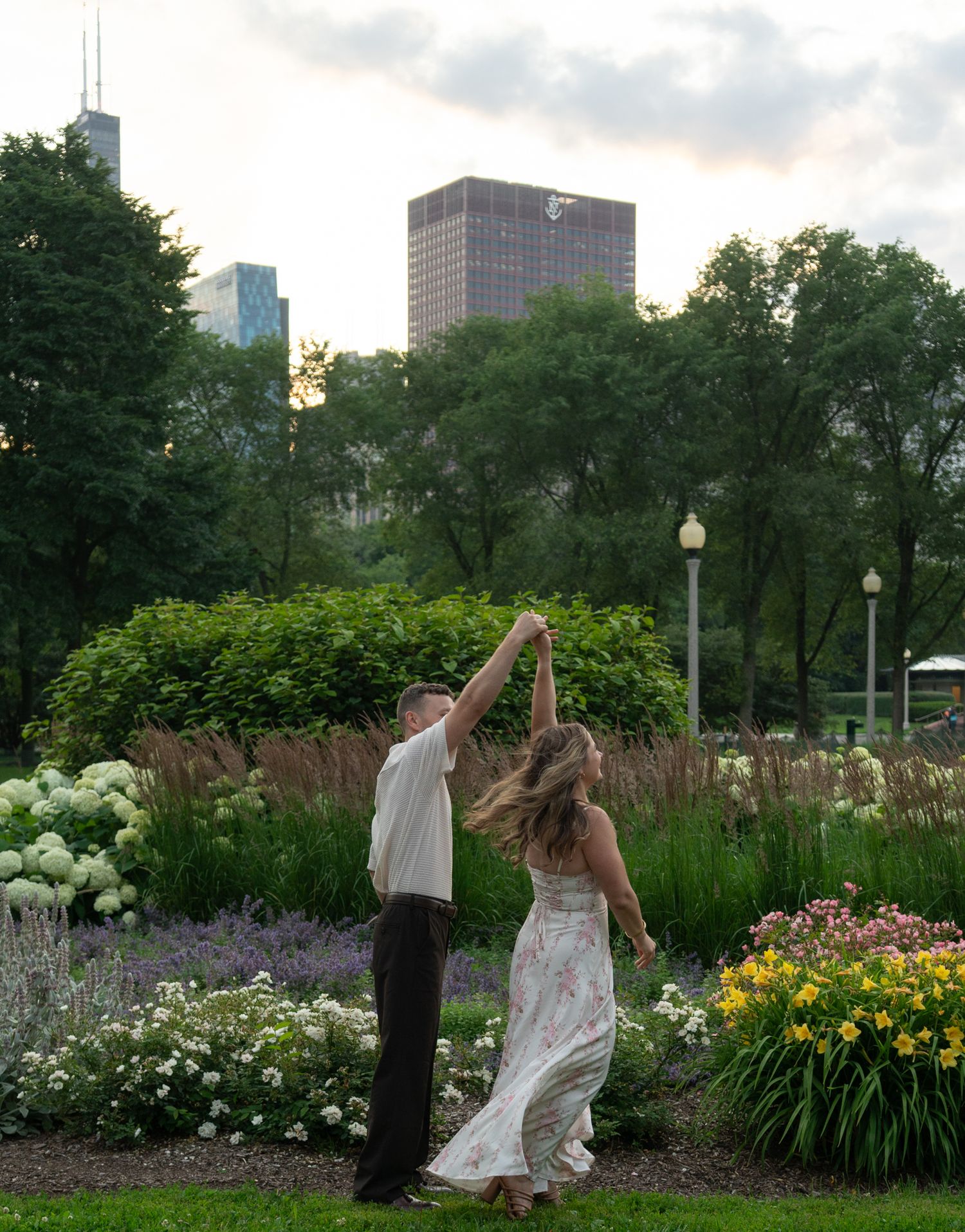 Couple dancing in a lush garden park with Chicago city skyline buildings visible through green trees at dusk.