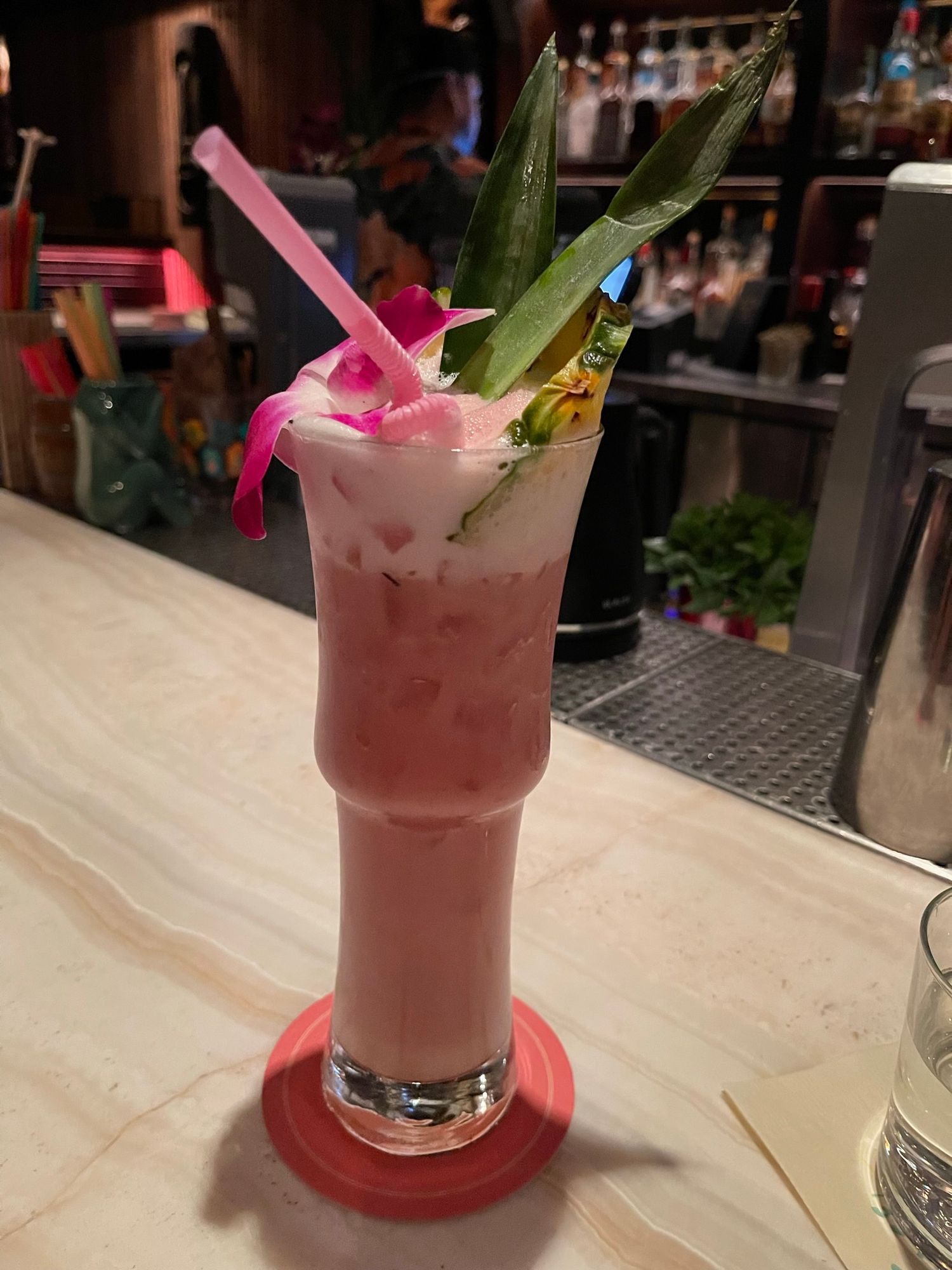 A tropical pink cocktail garnished with pineapple leaves and an orchid flower, served in a tall glass at a bar | Sydney Allen Photography