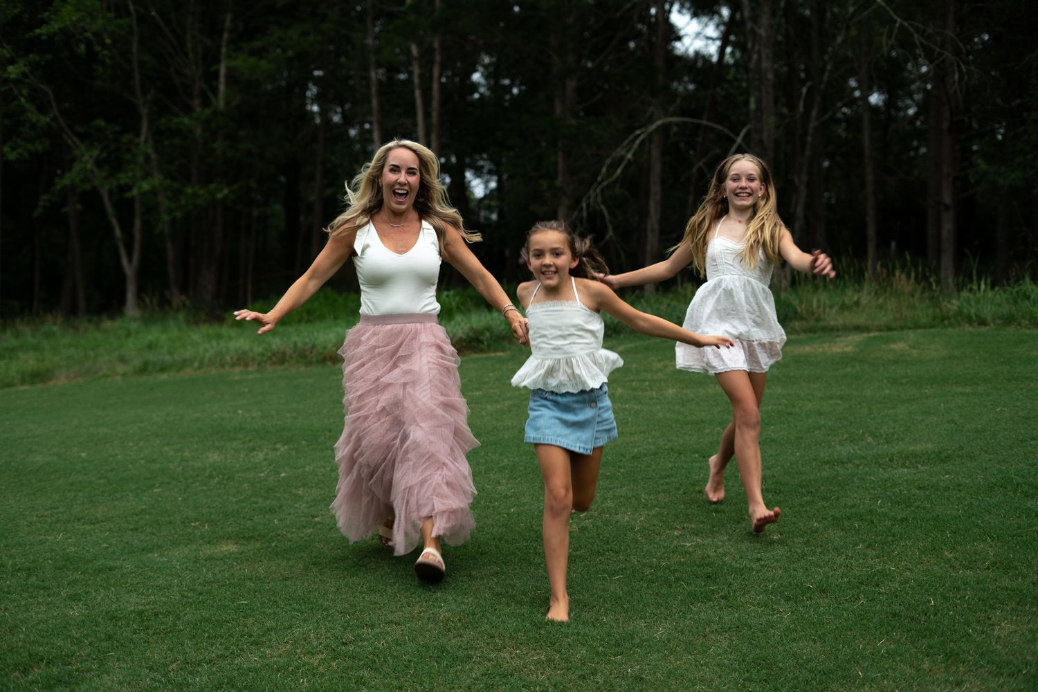 Woman and two girls run joyfully hand-in-hand across green lawn with forest backdrop.
