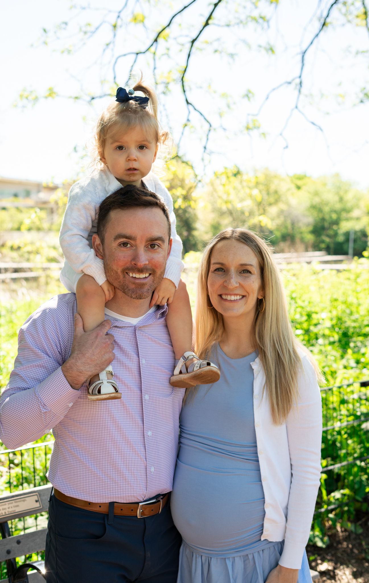 A smiling family of three poses outdoors, with a toddler girl sitting on dad's shoulders beside a pregnant mom.