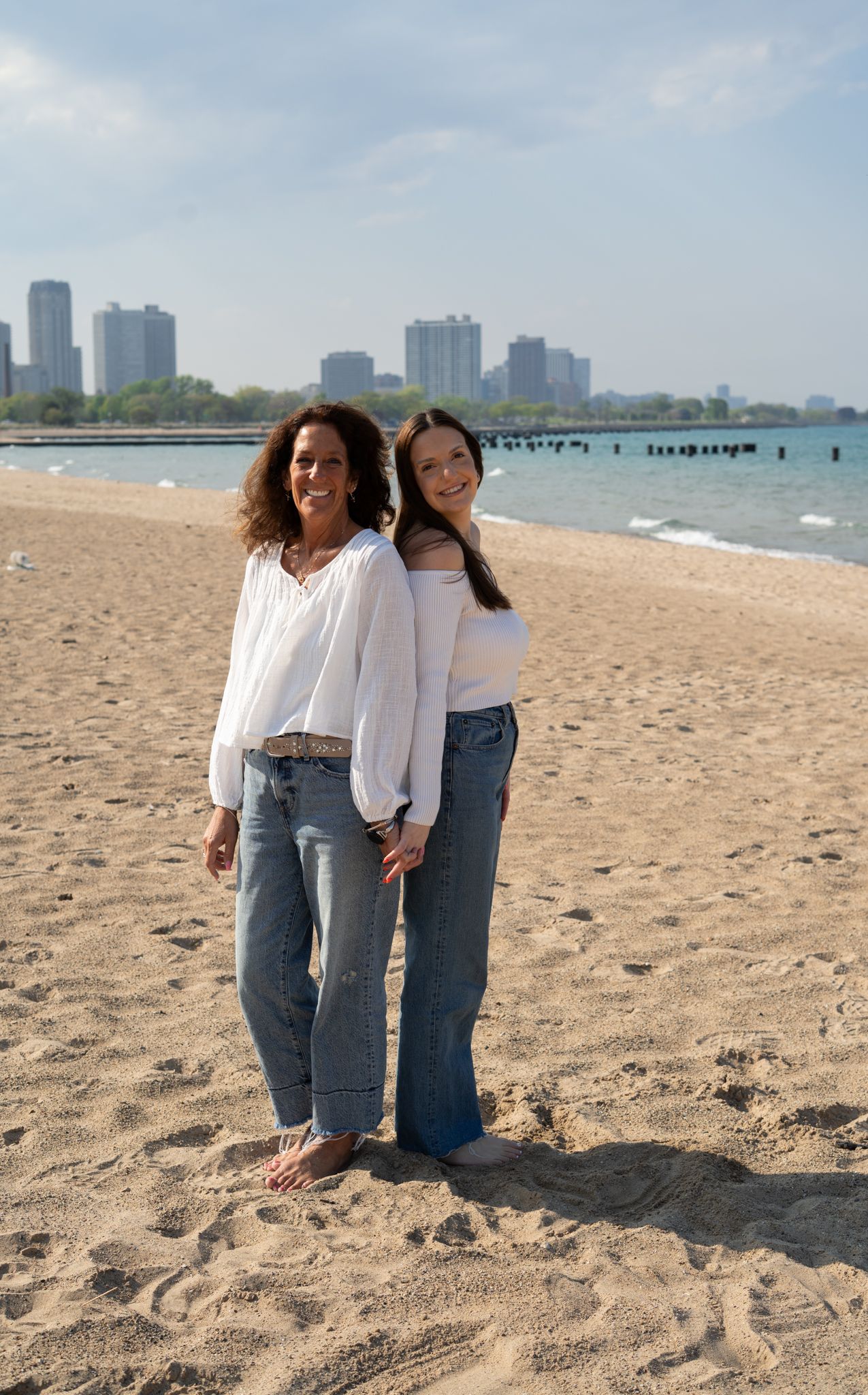 Two women in white tops and jeans smiling on a sandy beach with a city skyline in the background.